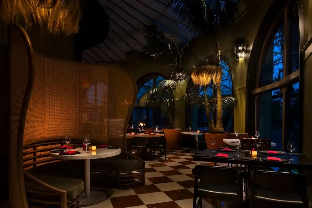 Dimly lit restaurant interior with curved booth seating, small tables set with red napkins and wine glasses, large potted plants, and arched windows showing a dark blue evening sky.