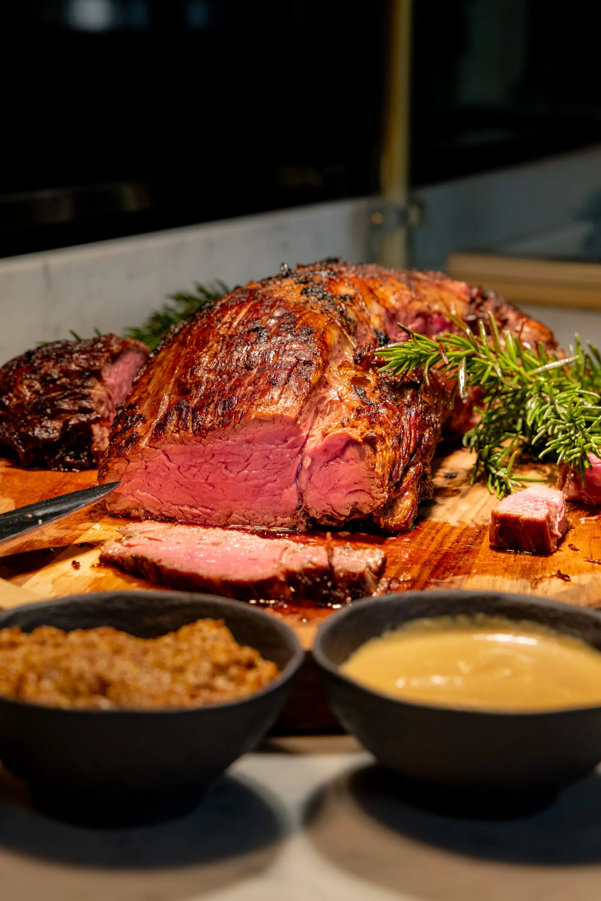 Sliced medium-rare roasted beef on a wooden cutting board garnished with rosemary, with two bowls of sauces in front.