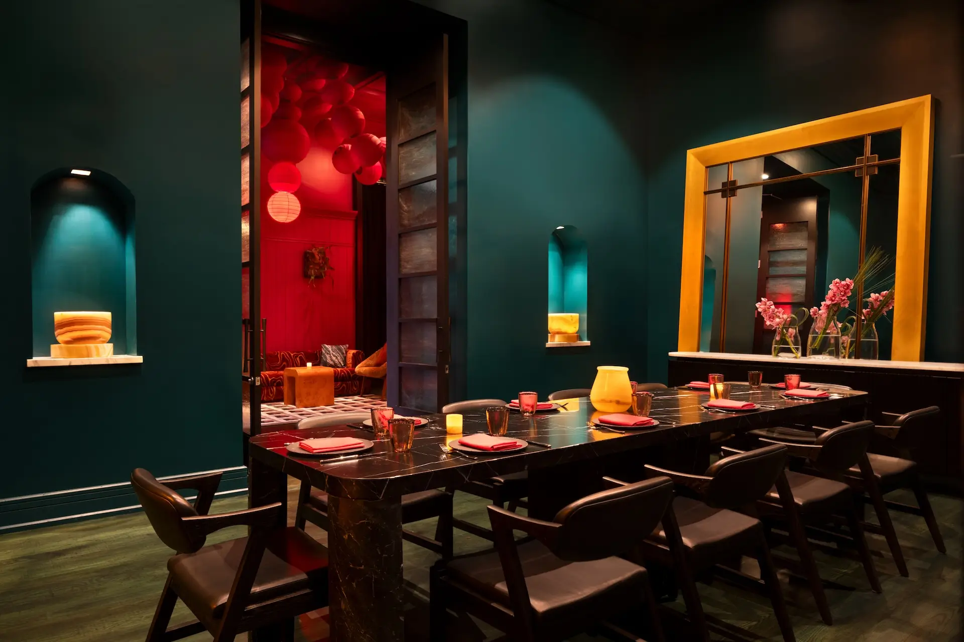 Elegant dining room with a long black marble table set for eight, dark chairs, teal walls, and a large yellow-framed mirror reflecting pink flowers.