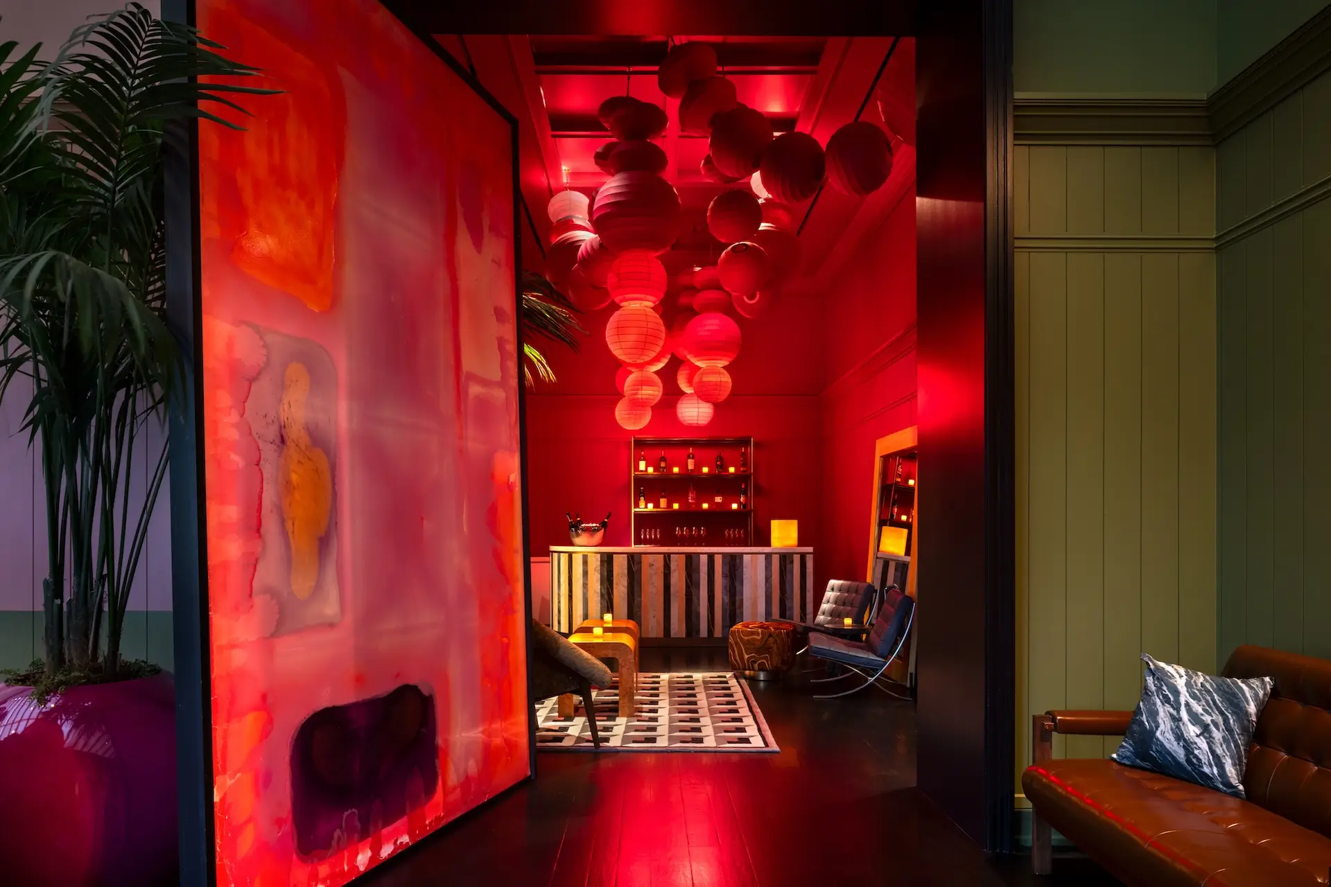 Dimly lit lounge area with a modern bar, hanging red lanterns, patterned rug, and cozy seating including a leather couch and chairs.