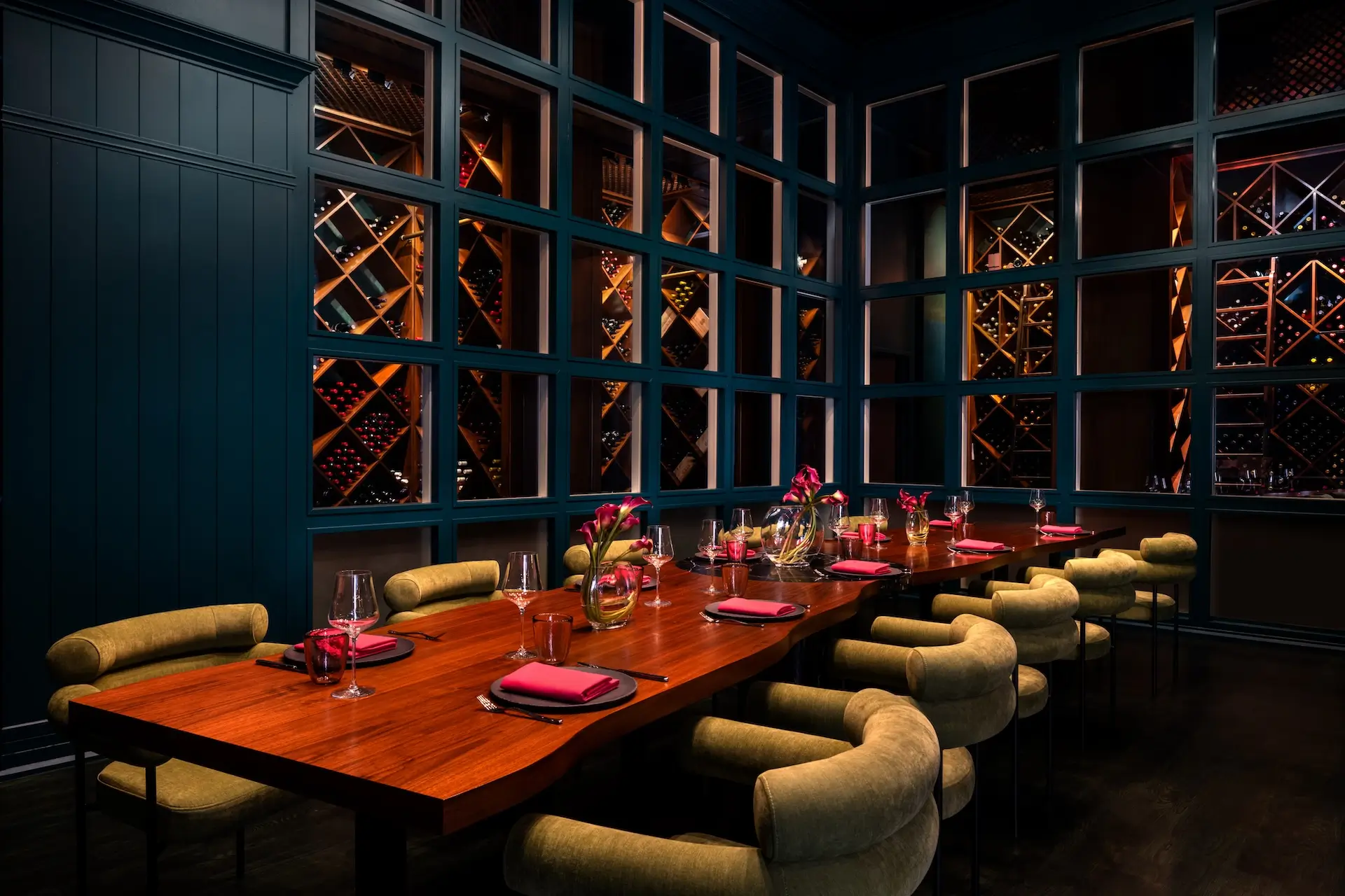 Elegant dining room with a large wooden table set with wine glasses, red napkins, and vases of pink flowers, surrounded by cushioned beige chairs, and a backdrop of wine racks behind glass panels.