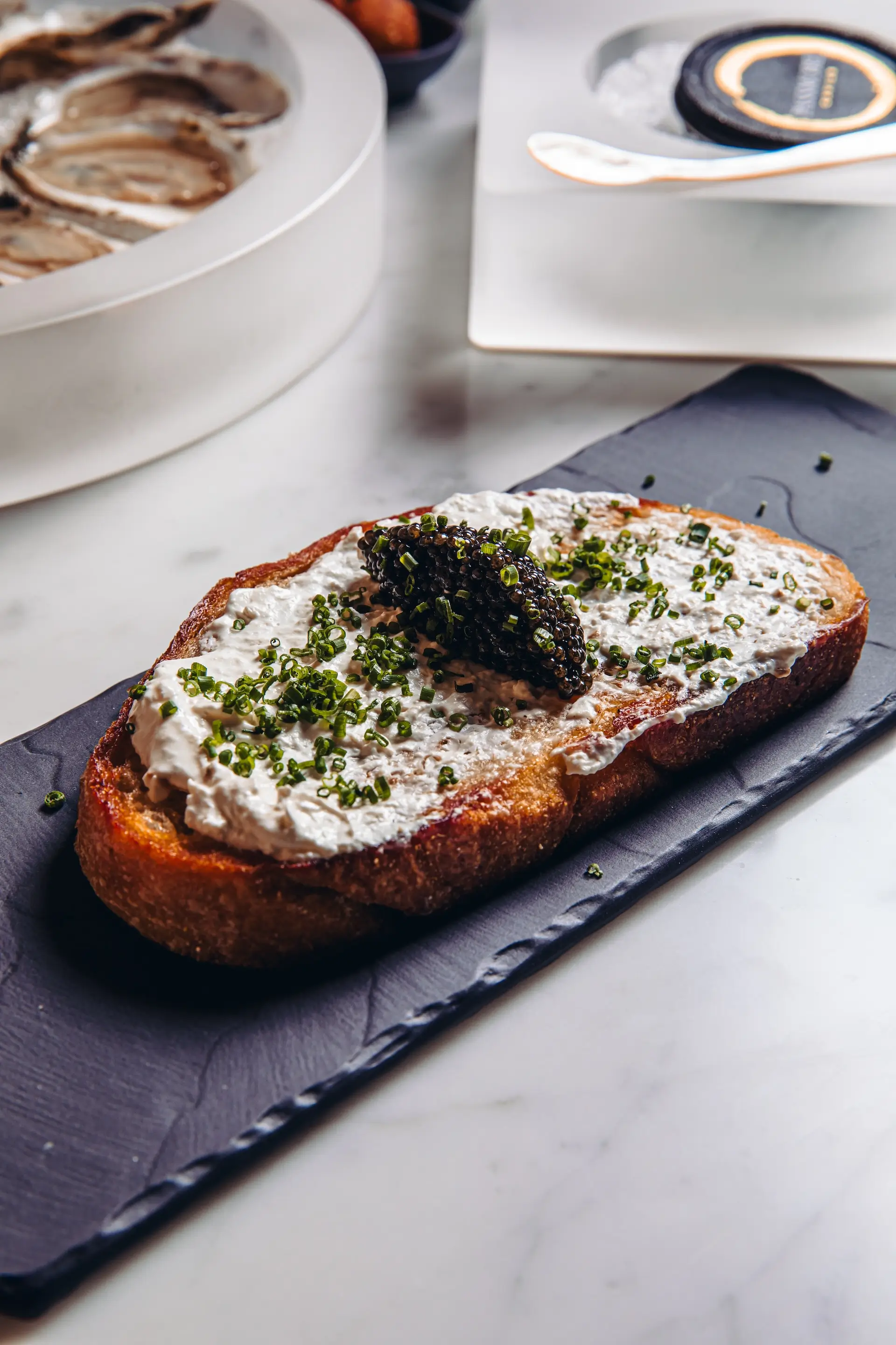 Slice of toasted bread topped with a creamy spread, a dollop of black caviar, and chopped chives on a black slate plate.