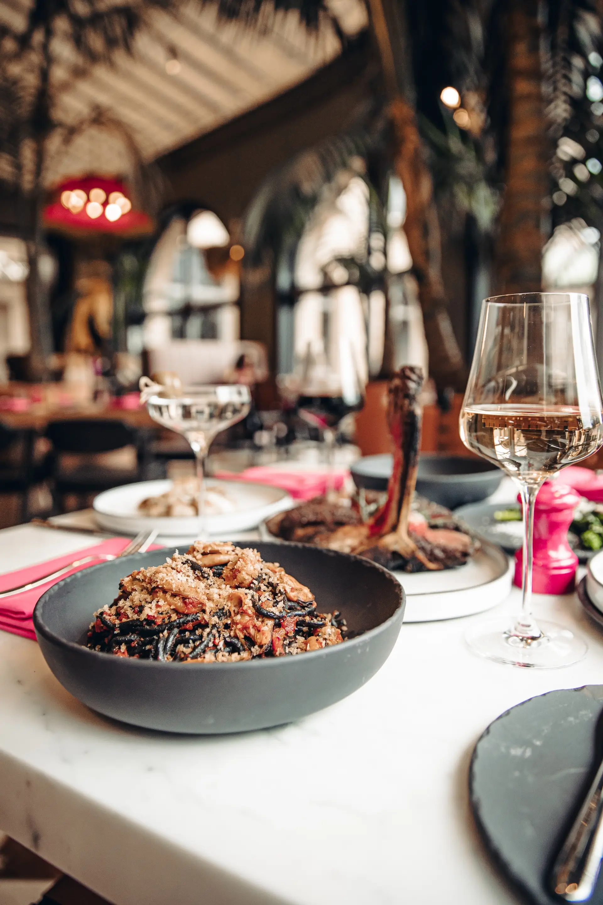 Black pasta with sauce in a black bowl next to a glass of white wine on a restaurant table.