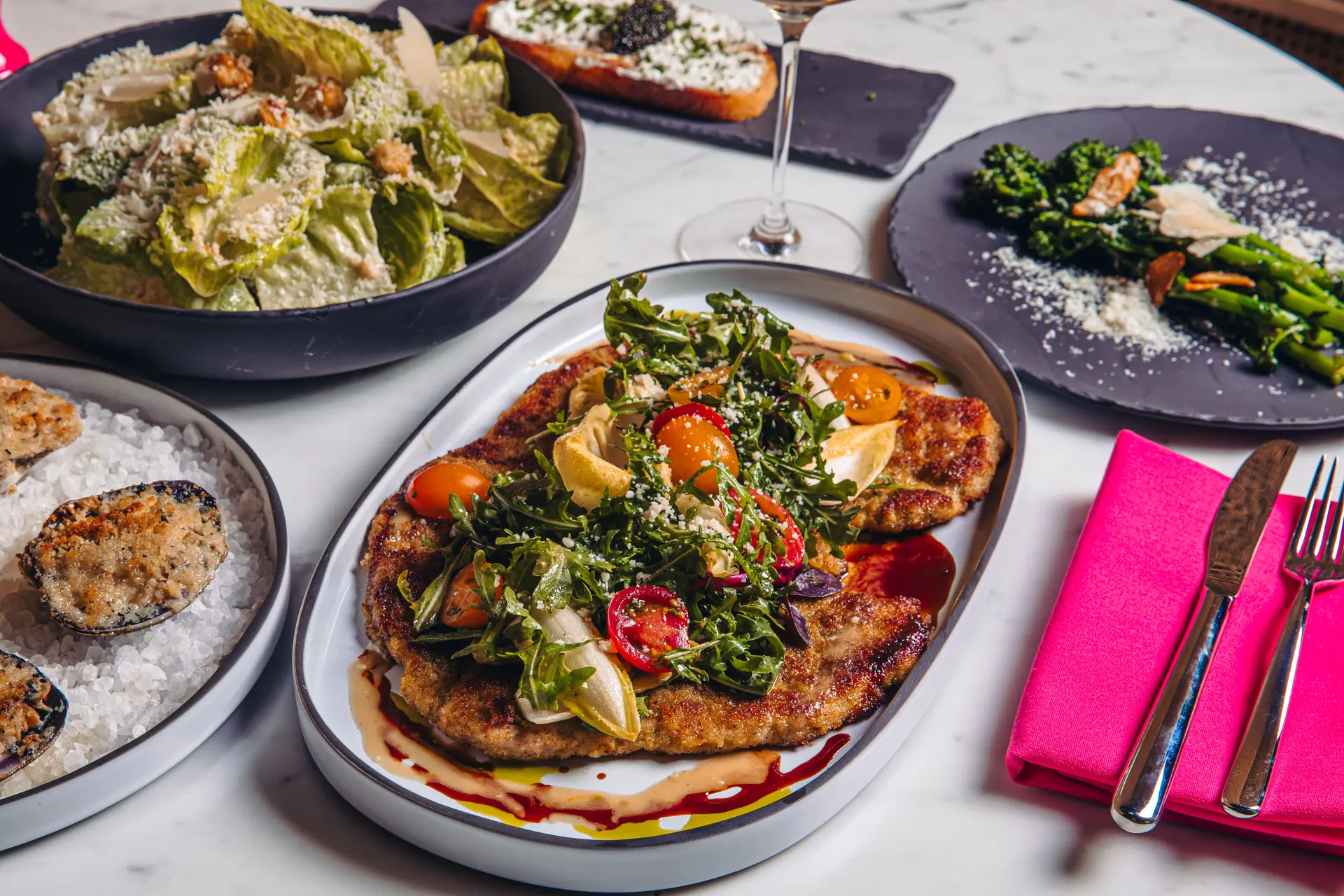 Breaded meat cutlet topped with mixed greens, cherry tomatoes, and lemon wedges on a white plate with sauce drizzles, surrounded by salad, stuffed mussels, sautéed greens, and a bread slice with cheese on a marble table with pink napkin and silver knife and fork.