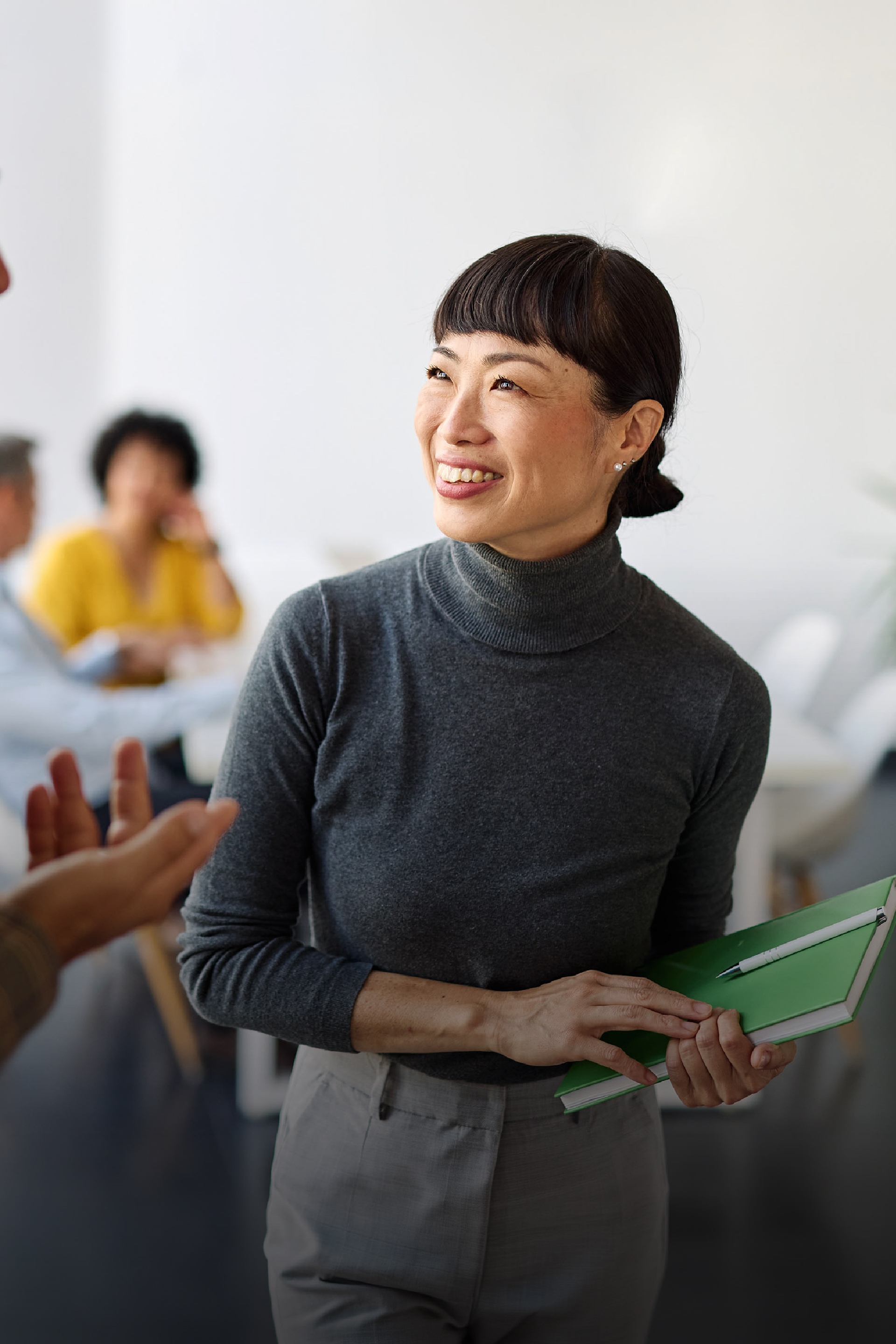 Smiling woman in a gray turtleneck and light pants holding a green notebook with a pen, engaging in conversation in a bright office setting with blurred coworkers in the background.