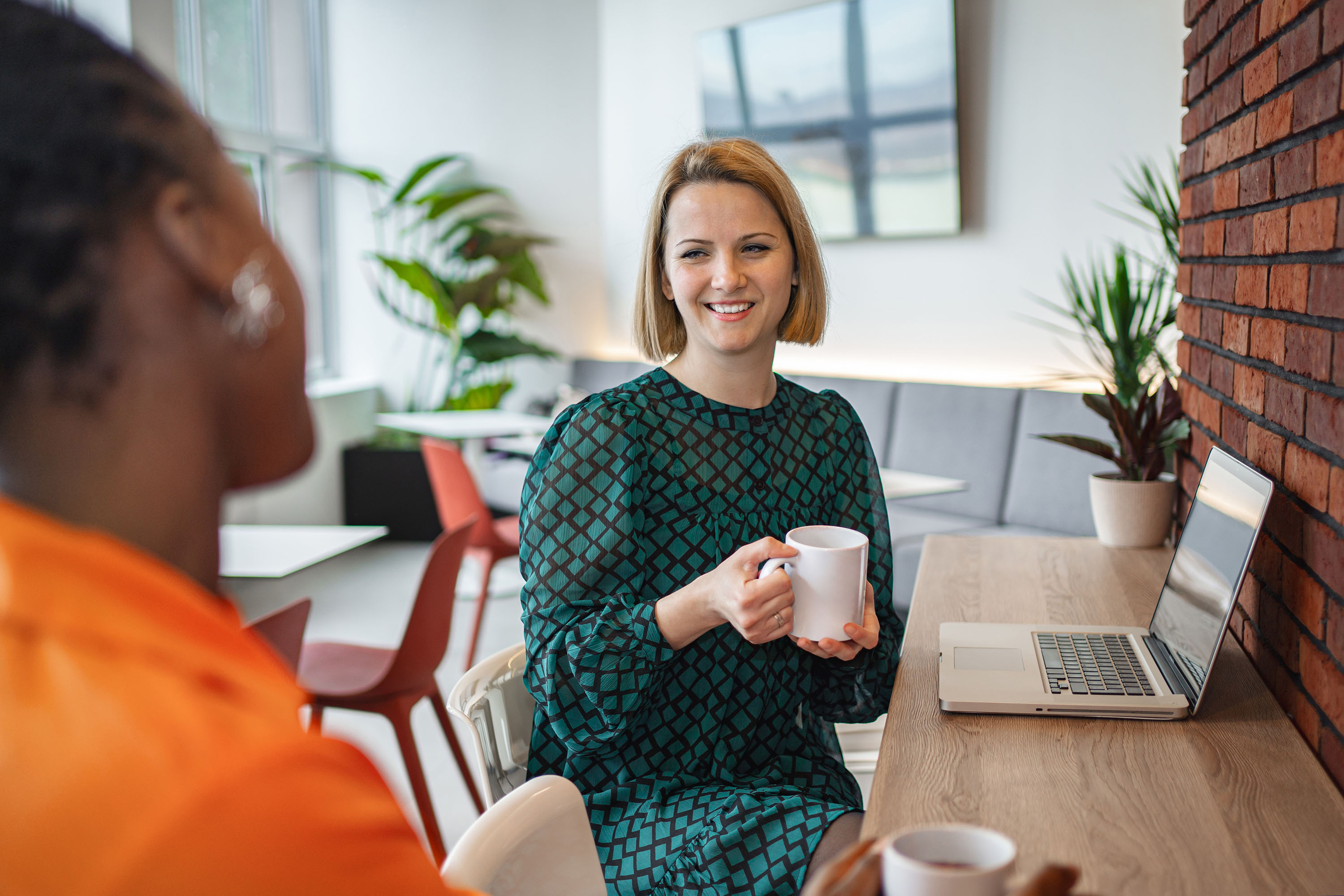 Two women sitting across from each other in a casual workspace, one smiling and holding a white mug near an open laptop on a wooden table, with plants and red chairs in the background.