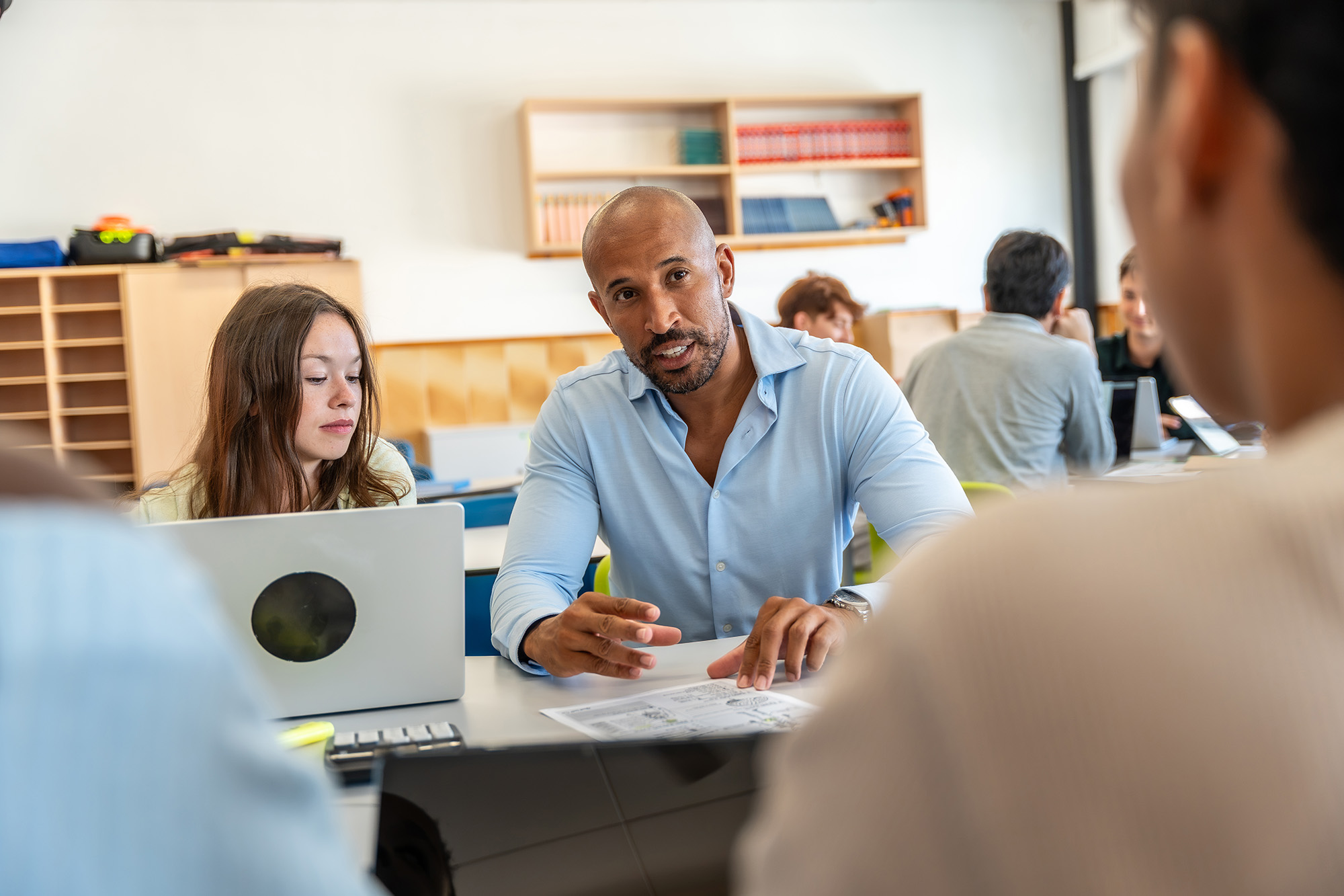 A man in a light blue shirt is engaging in a discussion while pointing at a document on a table, with a young woman next to him using a laptop and other people in the background in a classroom setting.