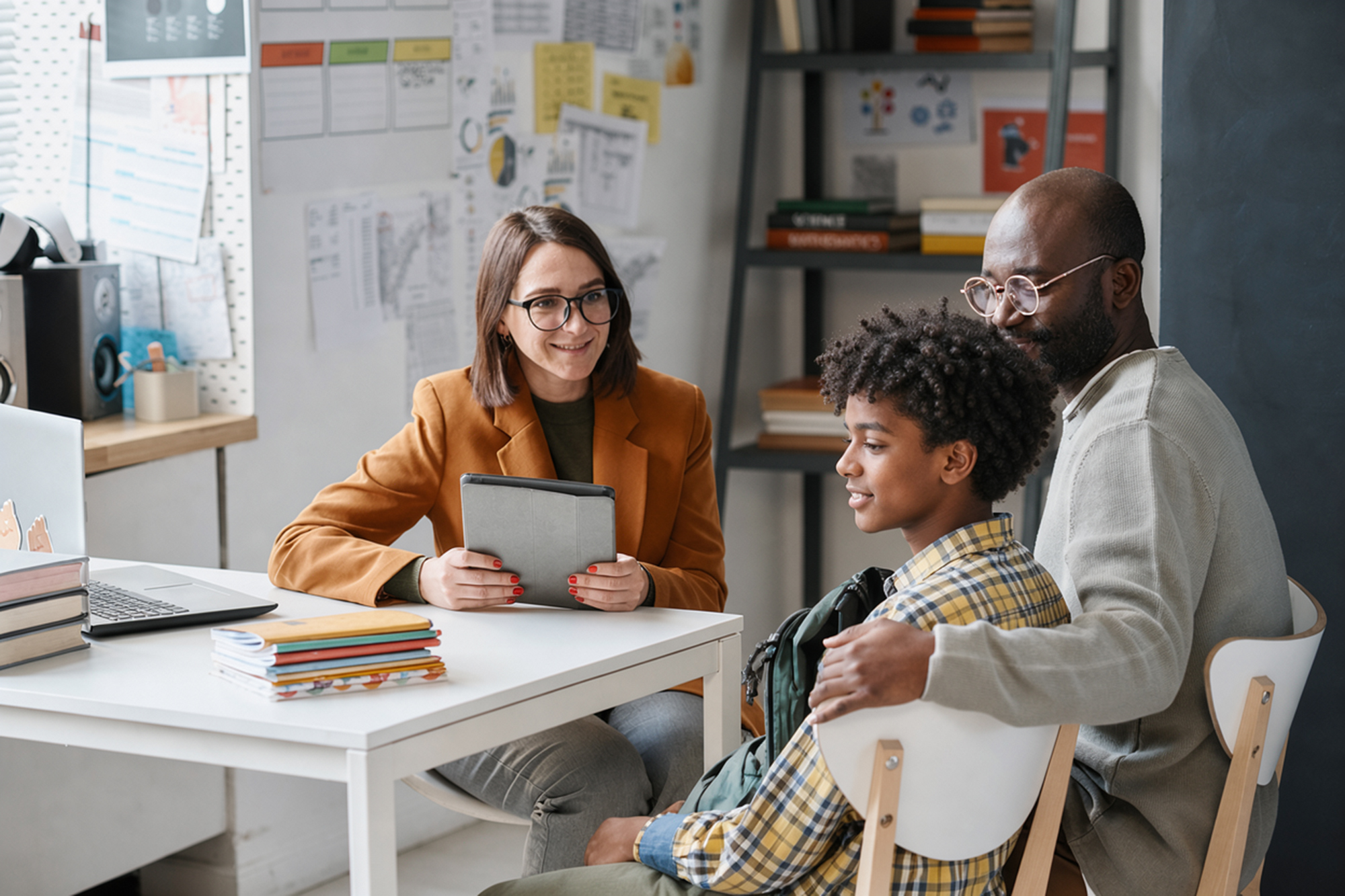 A female teacher in glasses and a brown jacket sits at a desk holding a tablet, talking to a teenage boy in a yellow plaid shirt and a man in a gray sweater seated next to him in a classroom.