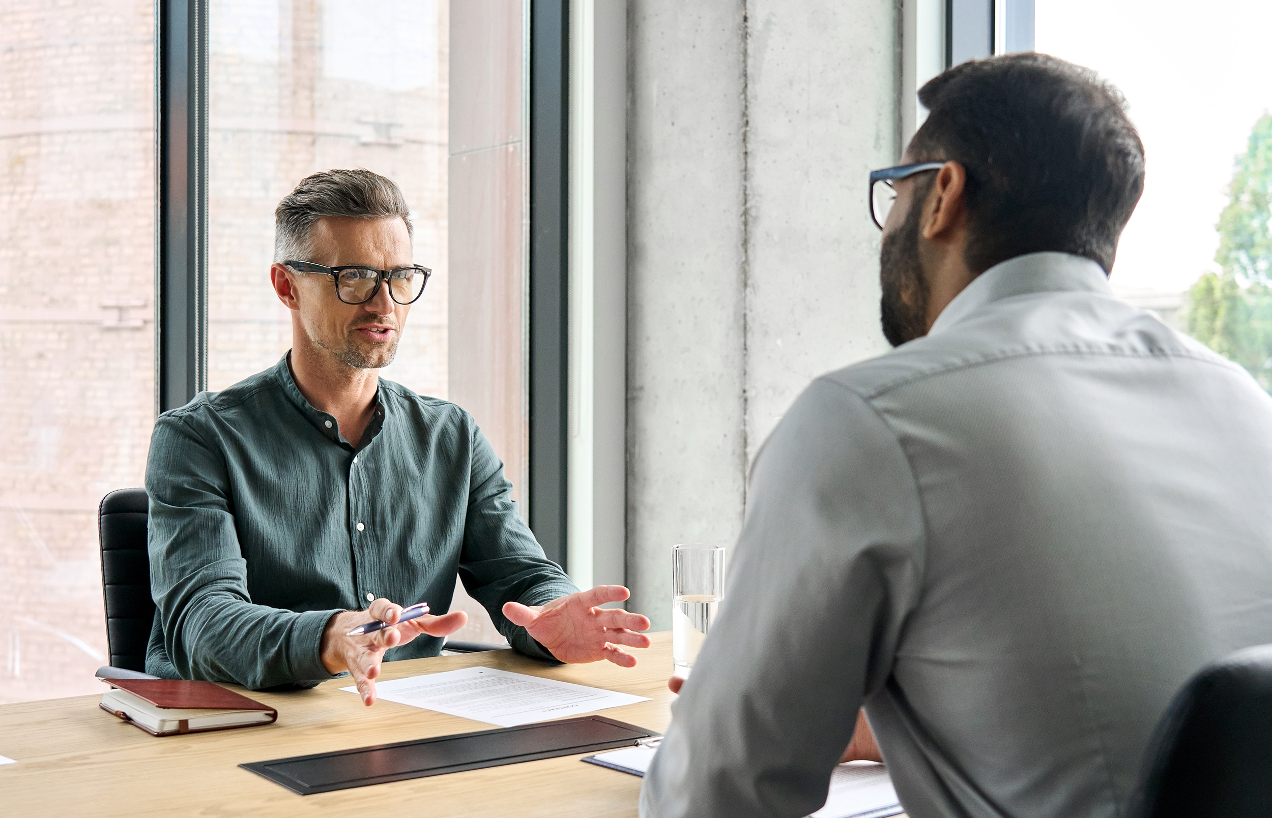 Two men wearing glasses engaged in a conversation across a wooden table in a modern office with large windows; one man gestures with his hands while holding a pen, and the other listens with a glass of water nearby.