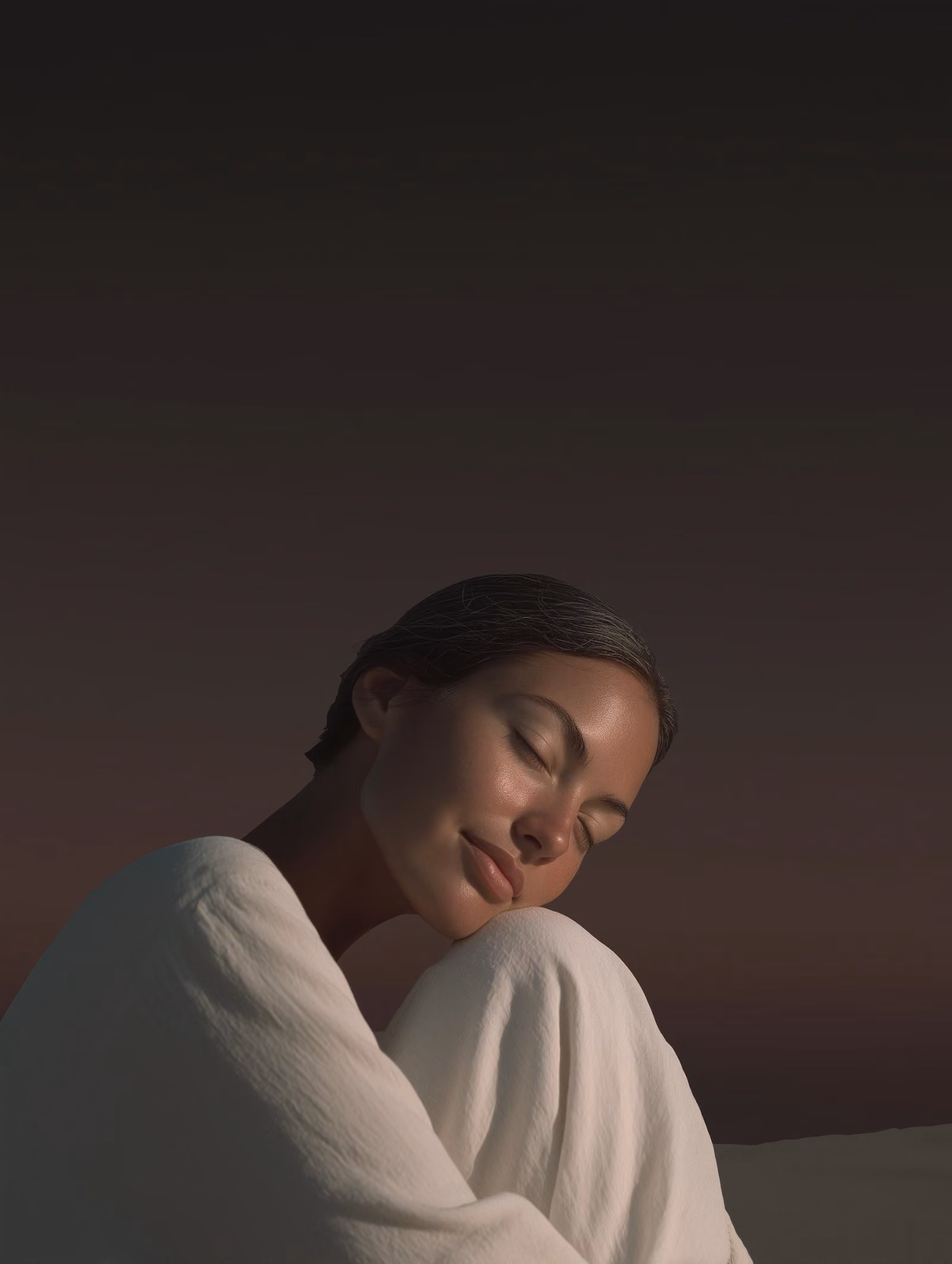 Woman with glowing, bare skin resting her head on her knees in soft daylight