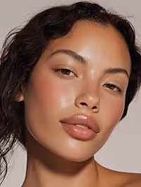 Close-up portrait of a woman with smooth, glowing skin, natural makeup, and dark curly hair against a plain neutral background.