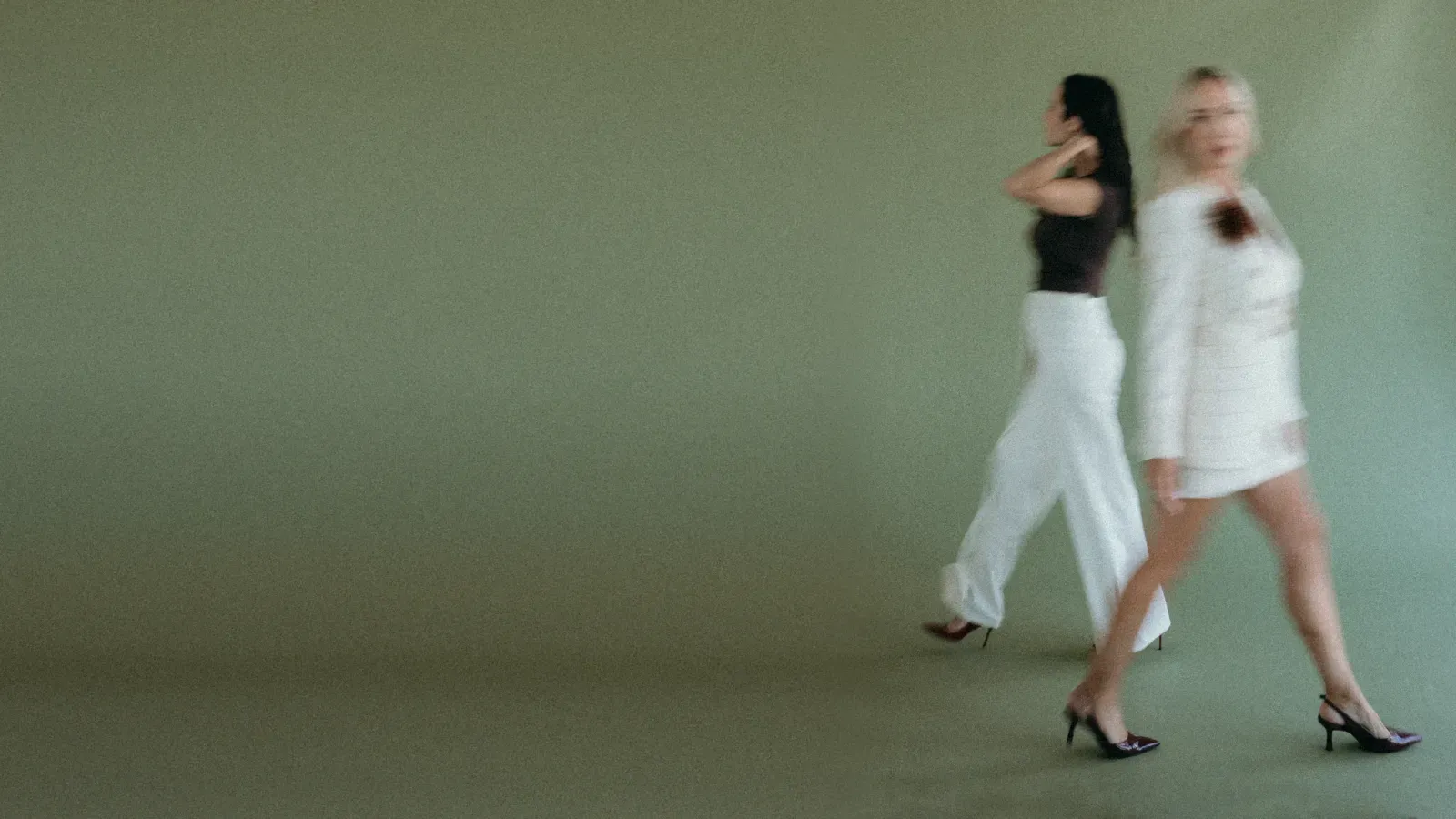Two women in tailored cream and black walking across a sage studio, softly blurred in motion.