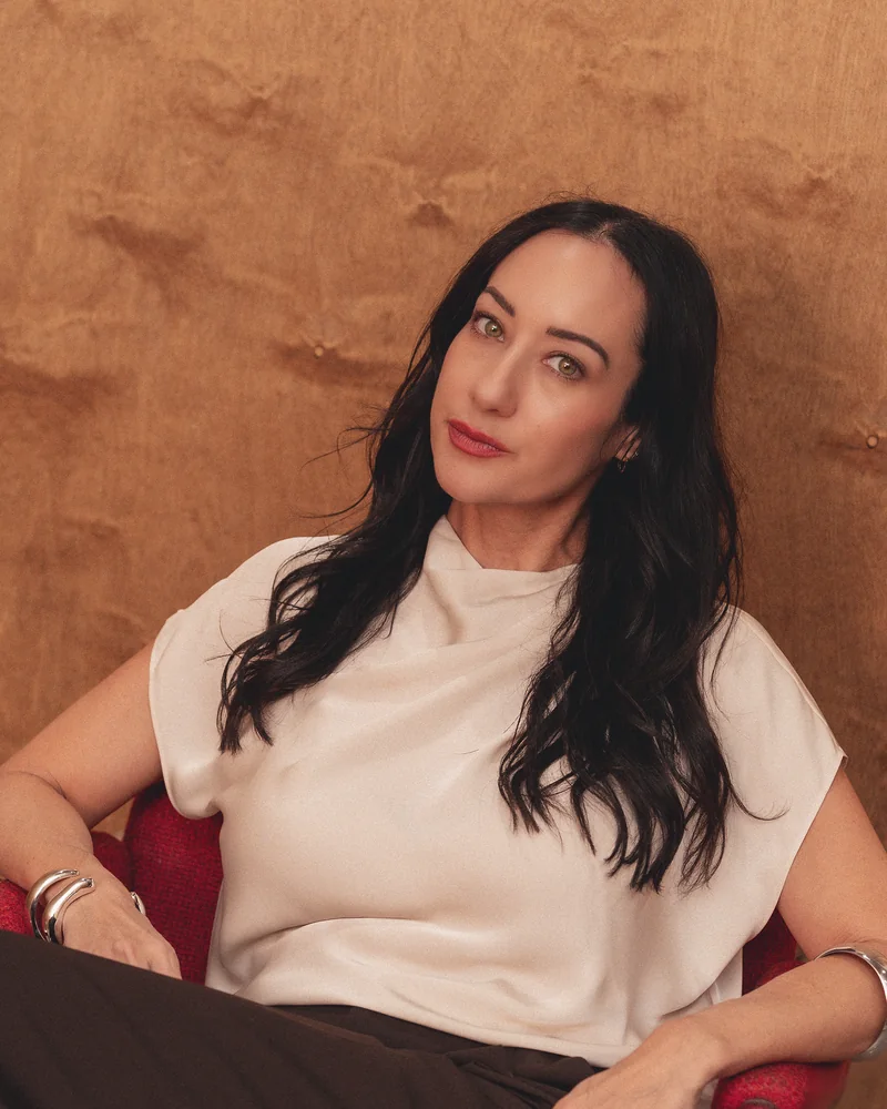 Jaclyn Freedman in a cream top, seated in a red chair against burl wood.