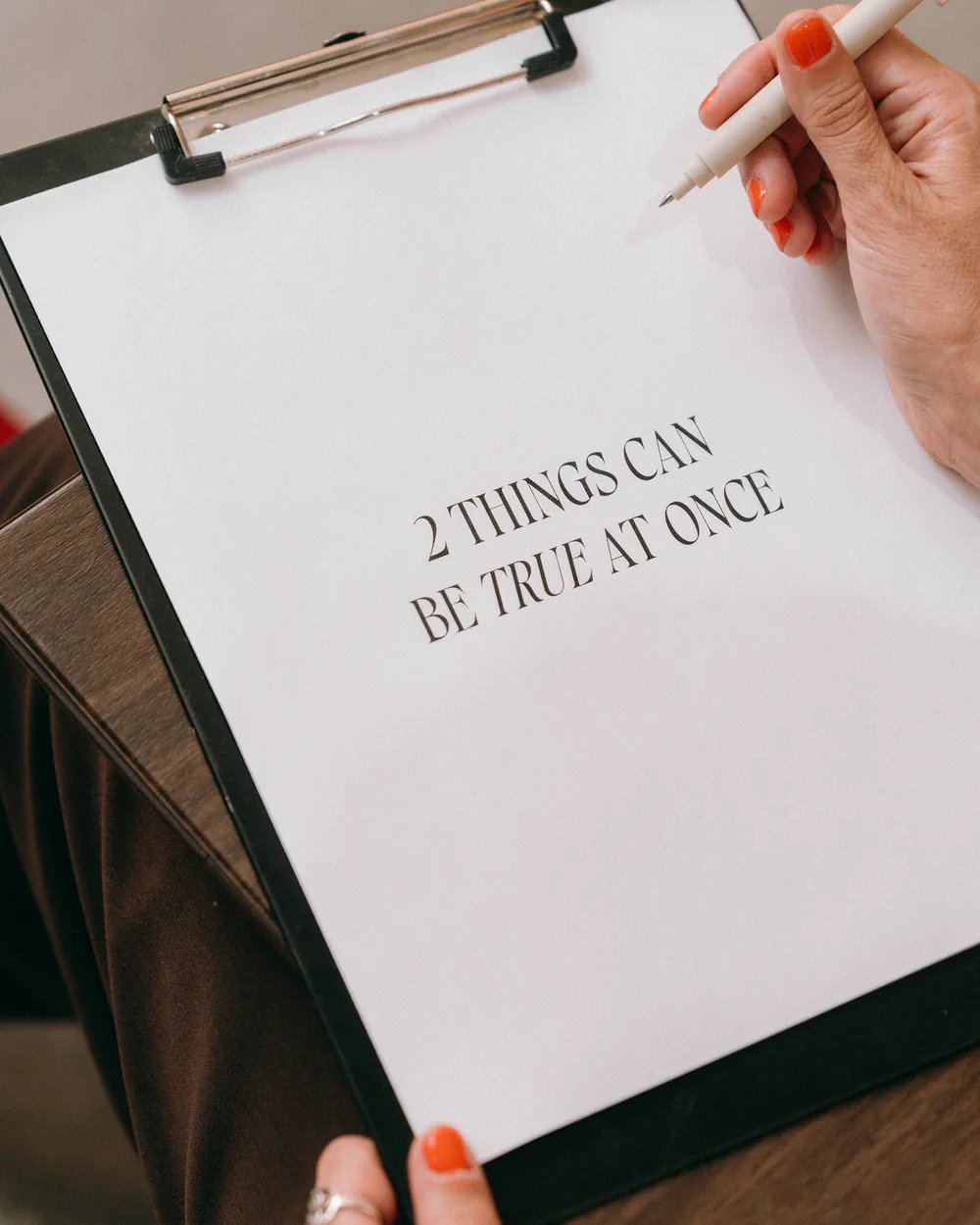A hand writing on a clipboard that reads "2 things can be true at once."