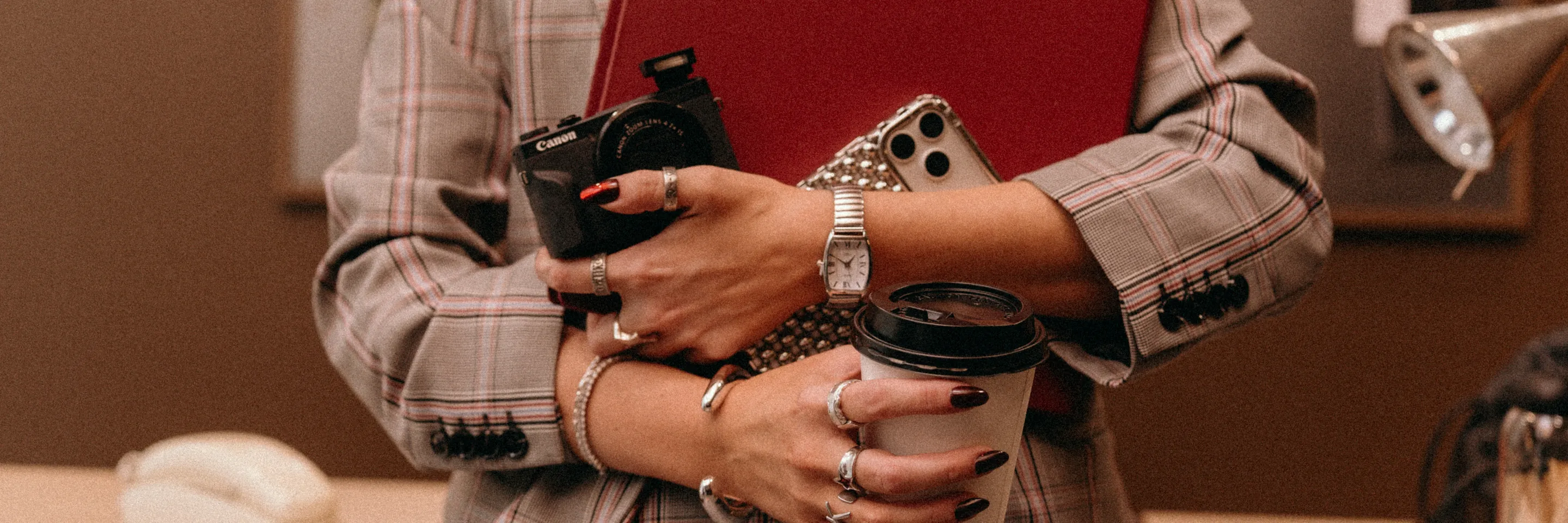 Hands holding a merlot binder, a camera, a phone and a coffee against a plaid blazer.