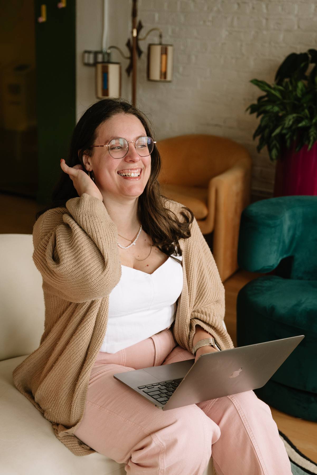 Smiling woman with glasses wearing a beige cardigan and pink pants seated on a white couch with a laptop on her lap in a cozy living room.