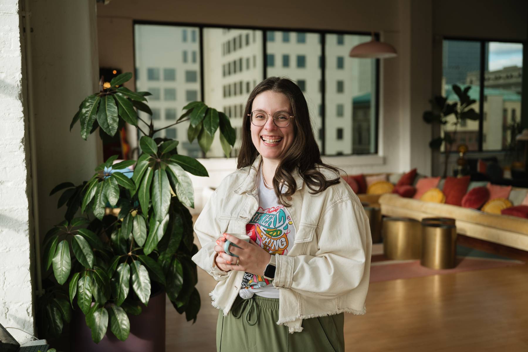 Smiling woman with long dark hair and glasses, wearing a white denim jacket and graphic t-shirt, holding a cup in a bright modern room with large windows, green potted plant, and colorful cushions on a beige couch.