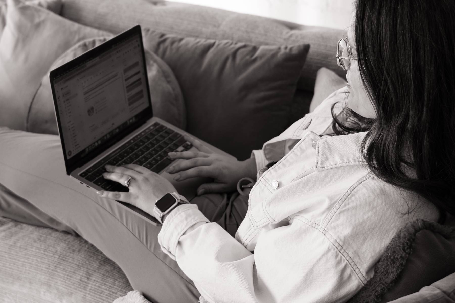 Person sitting on a couch using a laptop with an open email inbox, wearing glasses, a light-colored jacket, a smartwatch, and a ring on their left hand.