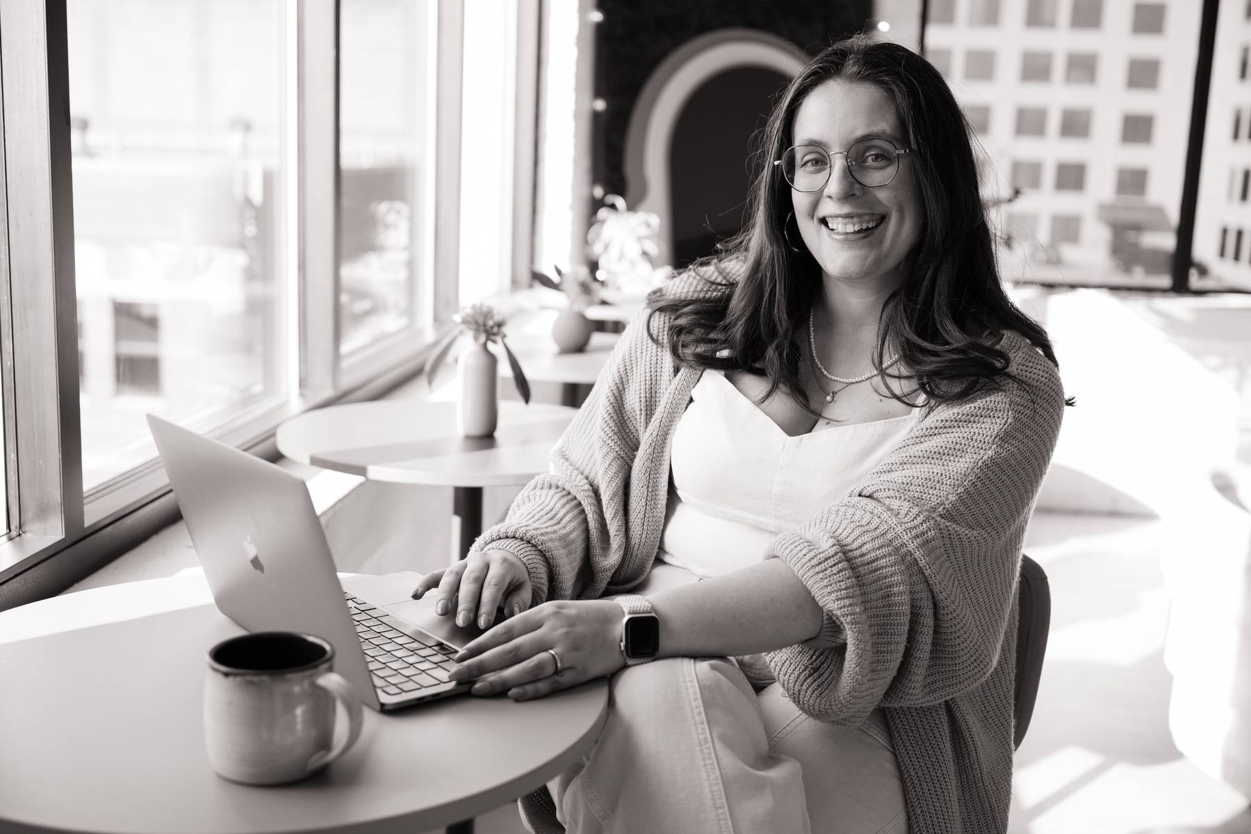 Molli smiling at her laptop at a café table by the window, a mug of coffee beside her