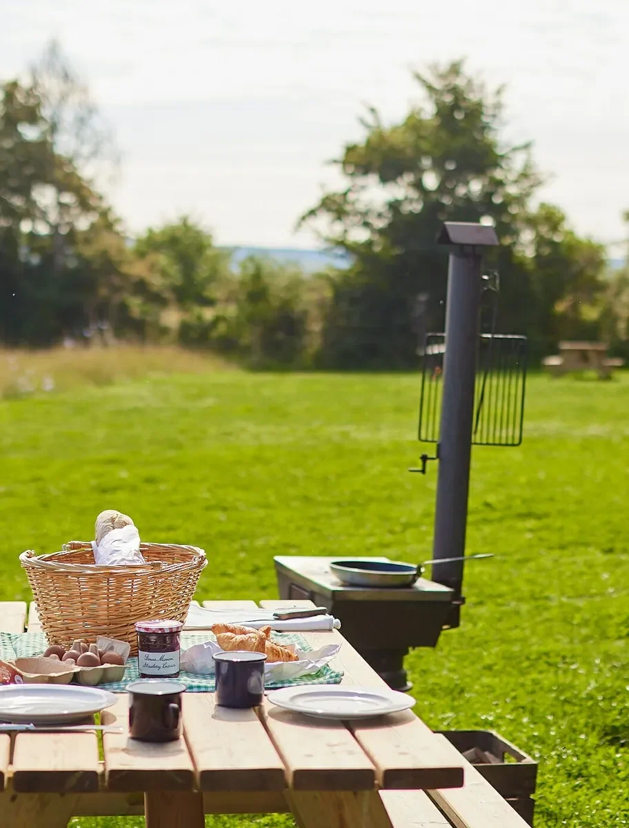 Picnic bench with a basket, cups and plates, a safari tent behind and open fields and trees in the distance