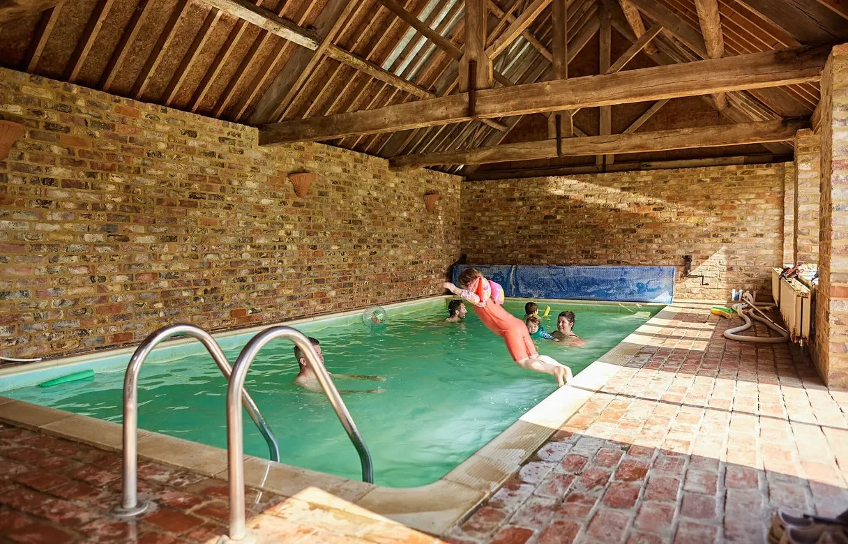 Swimming pool in an indoor barn with people in the pool and a child jumping in from the side