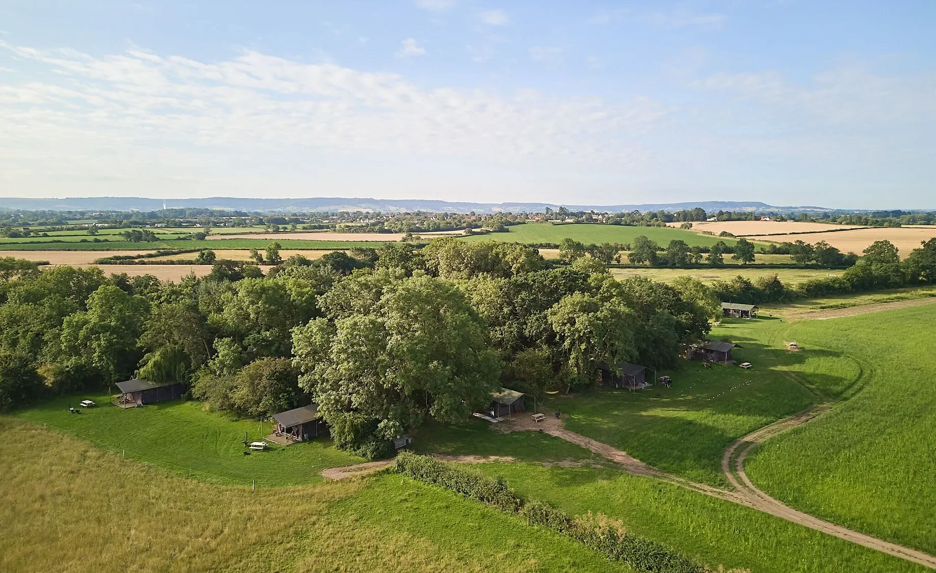 Aerial view of grass fields and clusters of trees, with glamping tents in a field and blue skies above