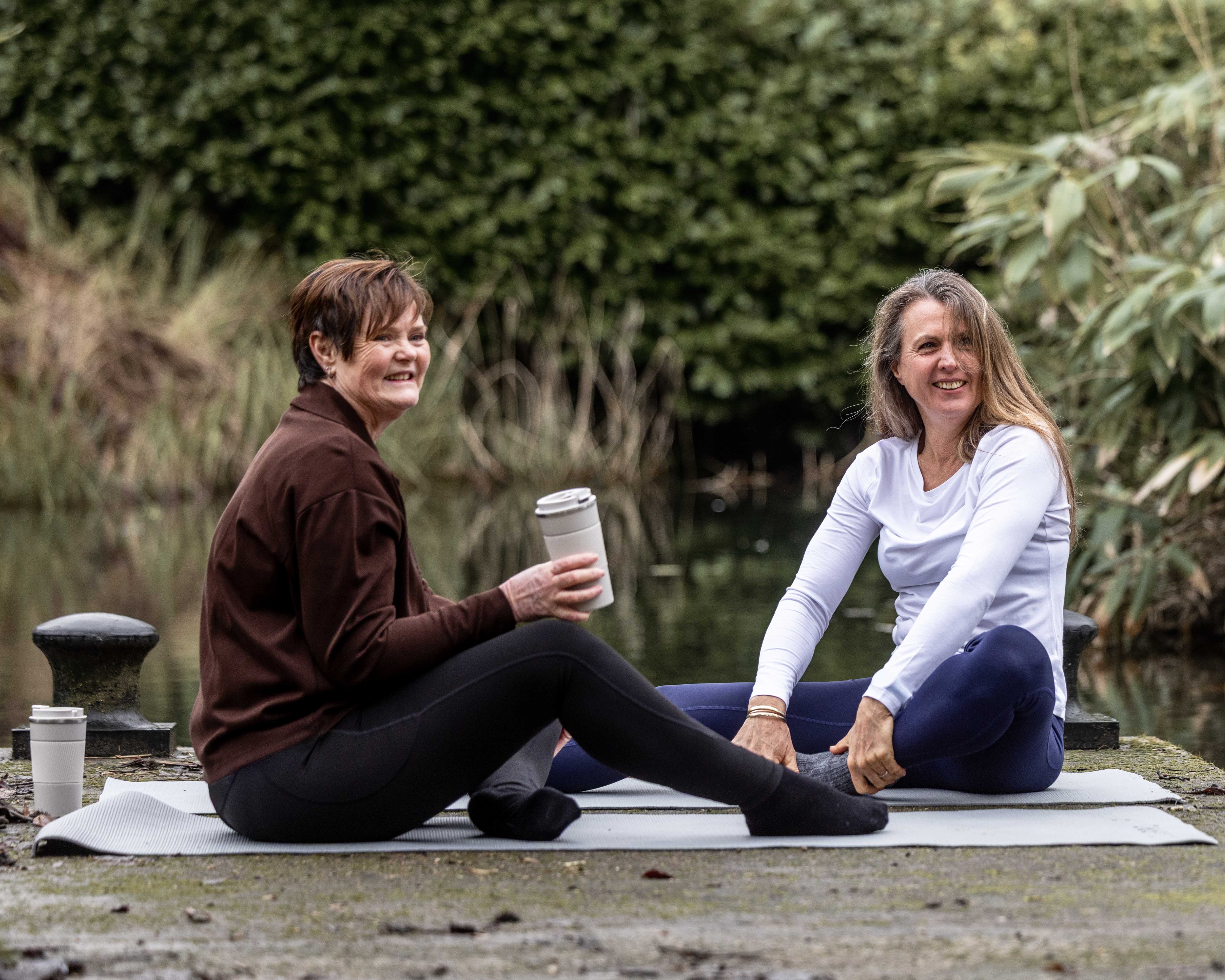 Two women practising yoga outdoors beside a lake