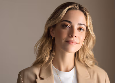 Blonde woman with light makeup wearing a beige blazer and white top, smiling softly against a neutral background.