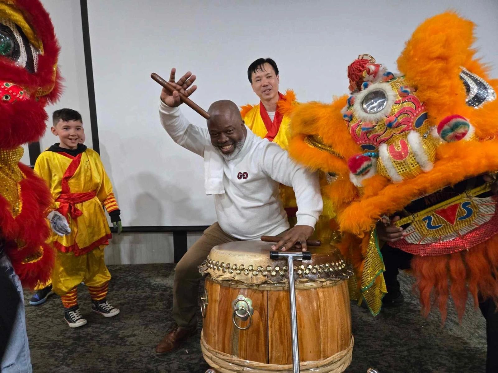 Sifu Bey playing the lion dance drum beside a young student and two lion dance costumes