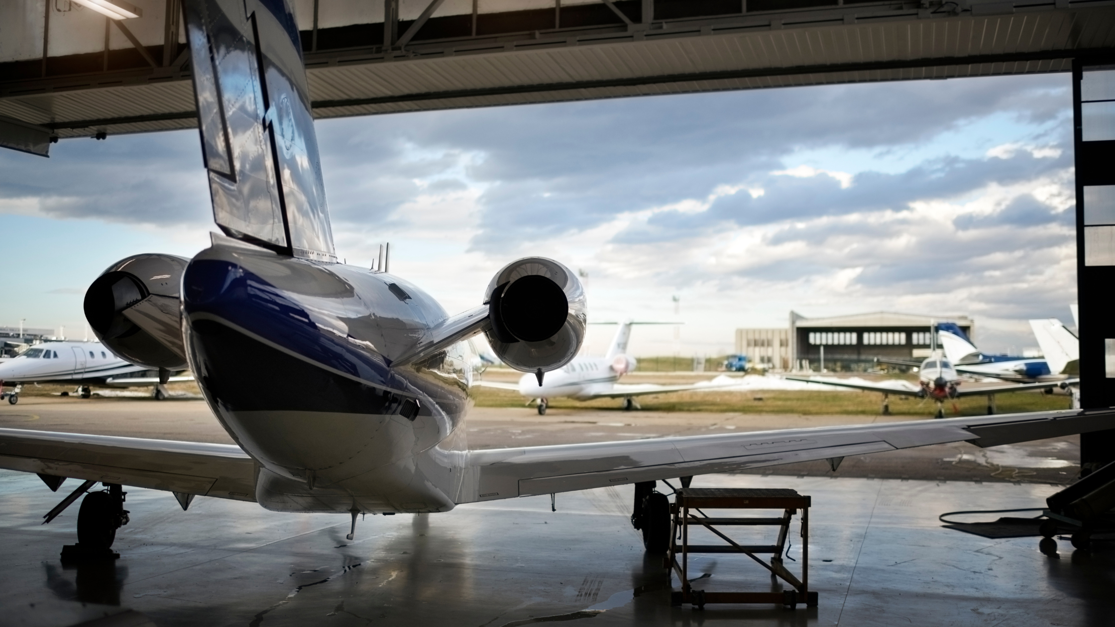 Private jet parked inside an aircraft hangar with other small planes visible outside under a cloudy sky.
