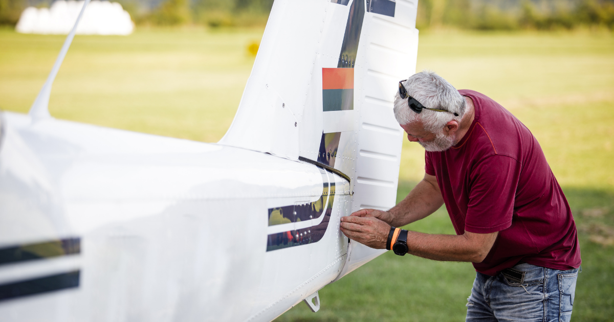 Man inspecting the tail section of a small white airplane on a grassy airfield.