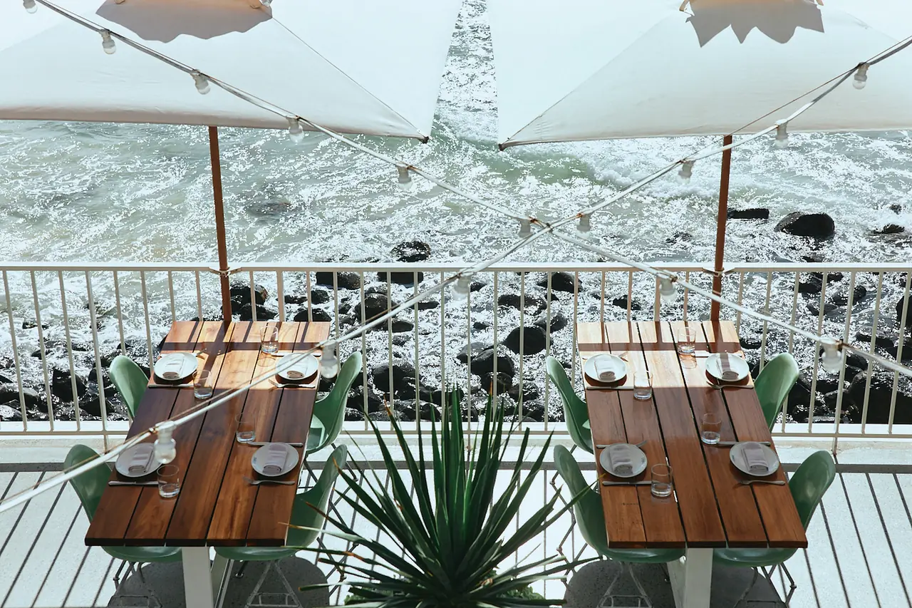 Set tables under white umbrellas above the rocks and surf at The Tropic