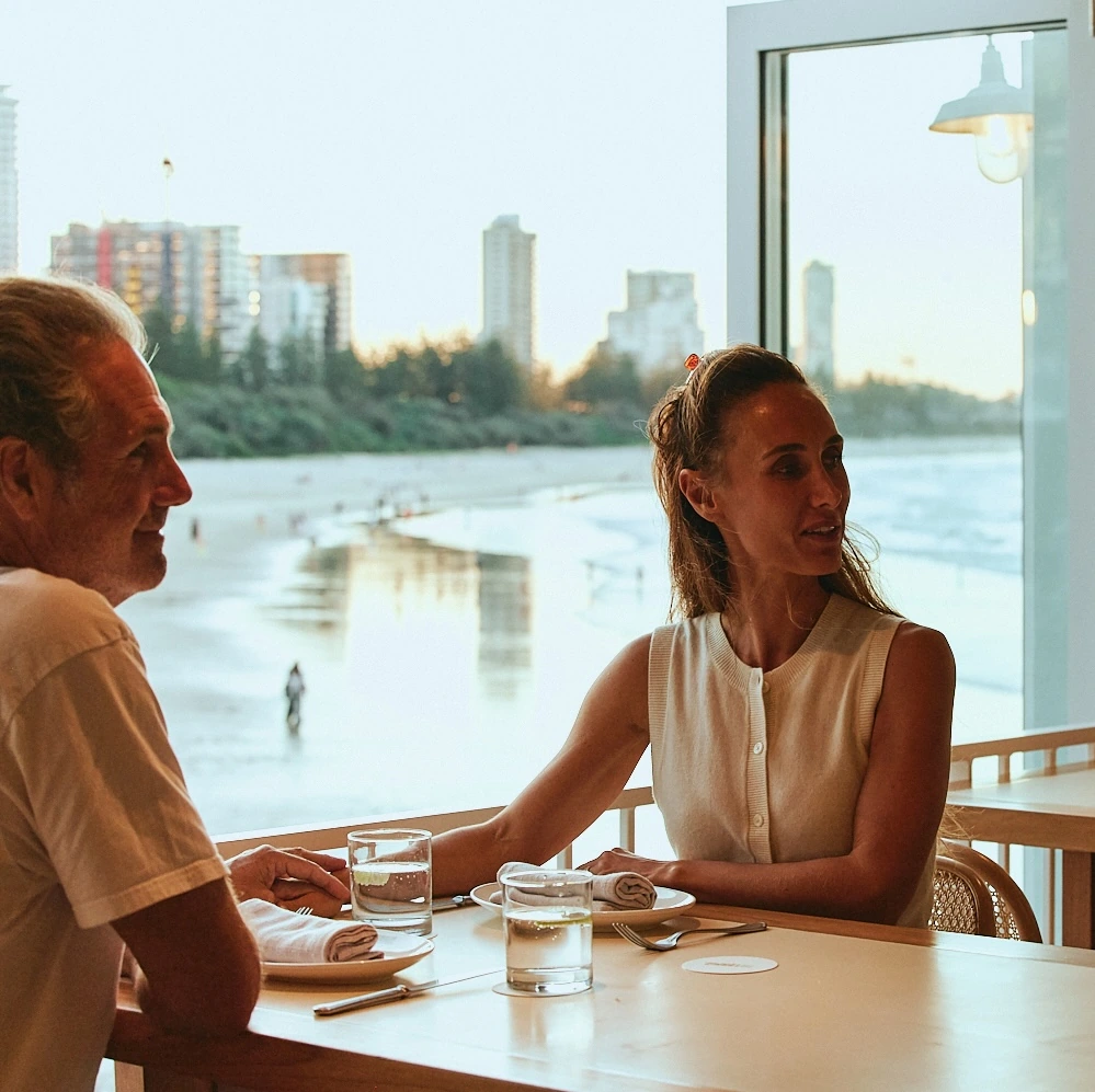 Couple dining by the window at The Tropic at dusk, with Burleigh Beach behind