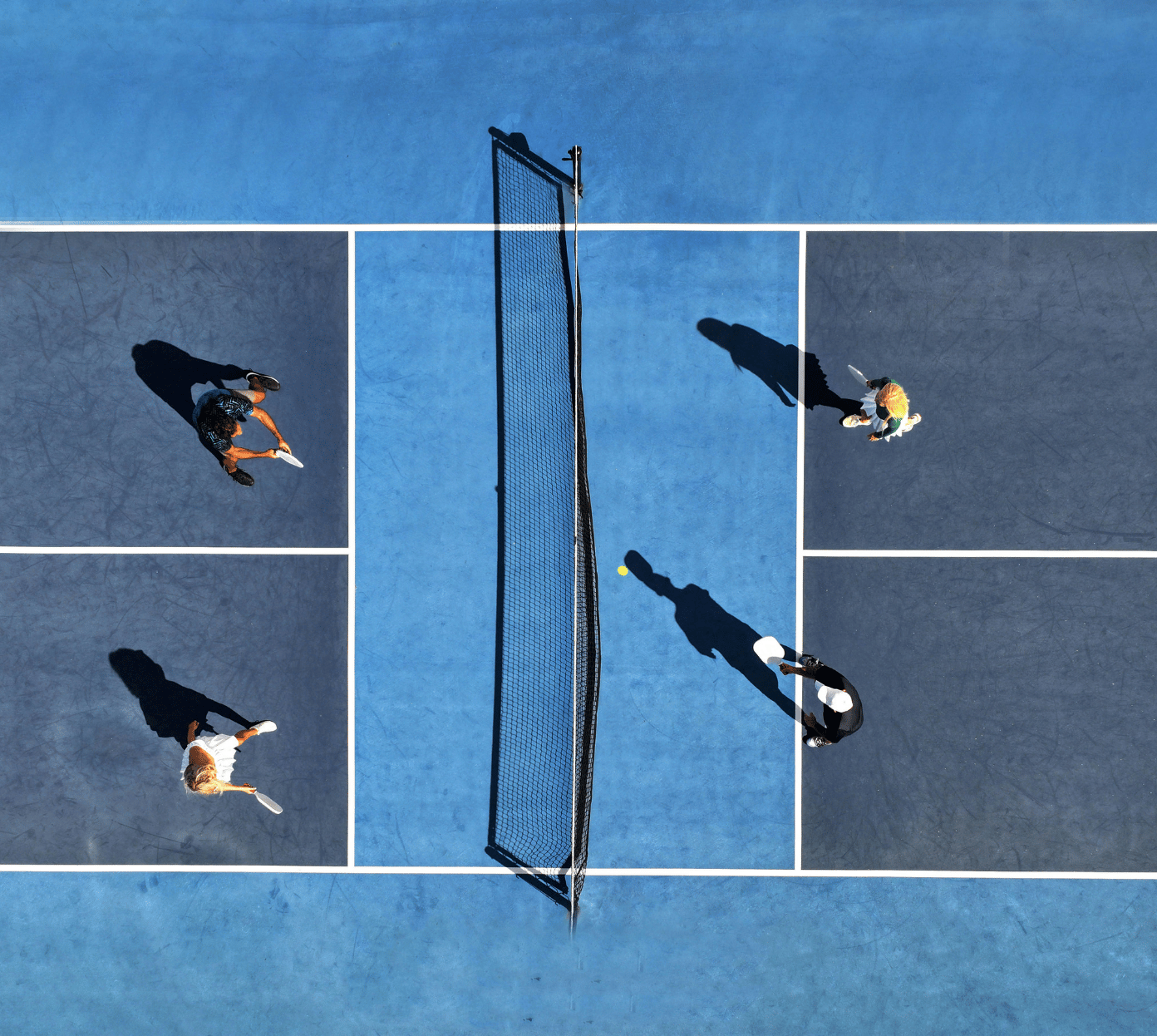 Aerial view of four people playing doubles pickleball on a blue court divided by a net, with two players on each side holding paddles and a yellow ball near the net.