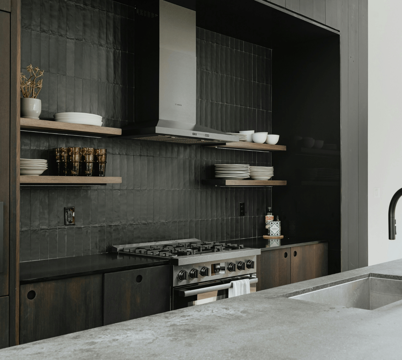 Modern kitchen with black tiled backsplash and dark wood cabinets, featuring a stainless steel gas stove with range hood, open wooden shelves holding white plates, bowls, and amber-colored glasses, and a light gray countertop with an inset sink and matte black faucet.