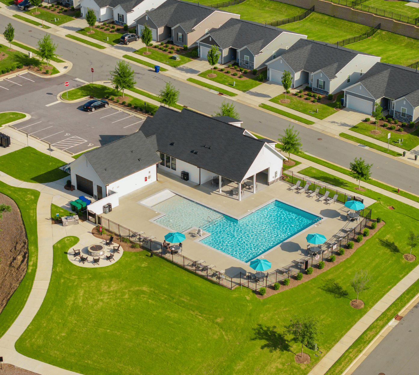 Aerial view of a residential community clubhouse featuring a large swimming pool with a smaller shallow pool adjacent, surrounded by a fenced concrete deck with lounge chairs, tables, and blue umbrellas. The clubhouse building has a covered patio with seating. Nearby is a circular fire pit area with chairs. Surrounding the clubhouse is well-maintained green lawn and a parking lot with cars. Rows of single-story houses line the opposite side of the street.