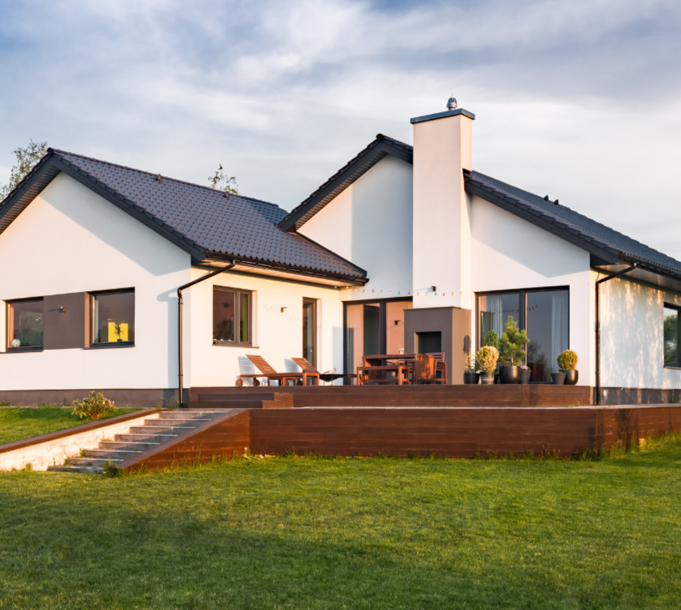 Modern single-story house with white exterior and dark grey roof, featuring large windows, a wooden patio with lounge chairs, a table, potted plants, and a grassy lawn under a partly cloudy sky.