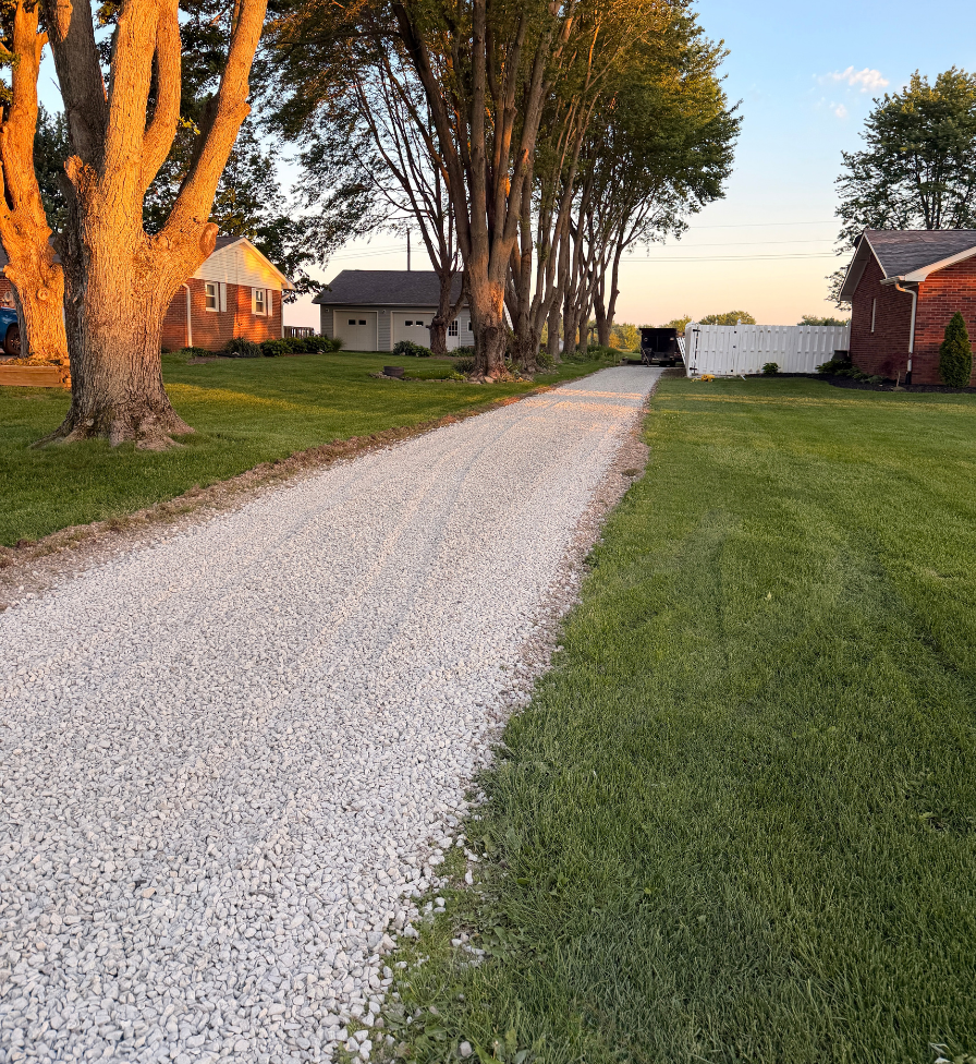 Gravel driveway runs between two green lawns lined with tall trees, leading to a garage and white fence under a clear sky at sunset.