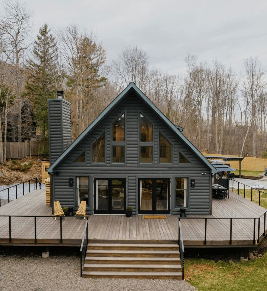 Modern dark gray log cabin with large triangular windows and wrap-around wooden deck featuring two chairs and a staircase with railing leading to gravel and grass.
