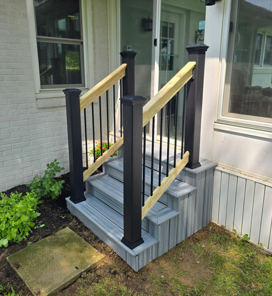 Small outdoor composite stairs with three steps and gray wood-like planks, featuring black metal posts and vertical balusters, light natural wood handrails, leading to a house entrance with white siding and large windows.