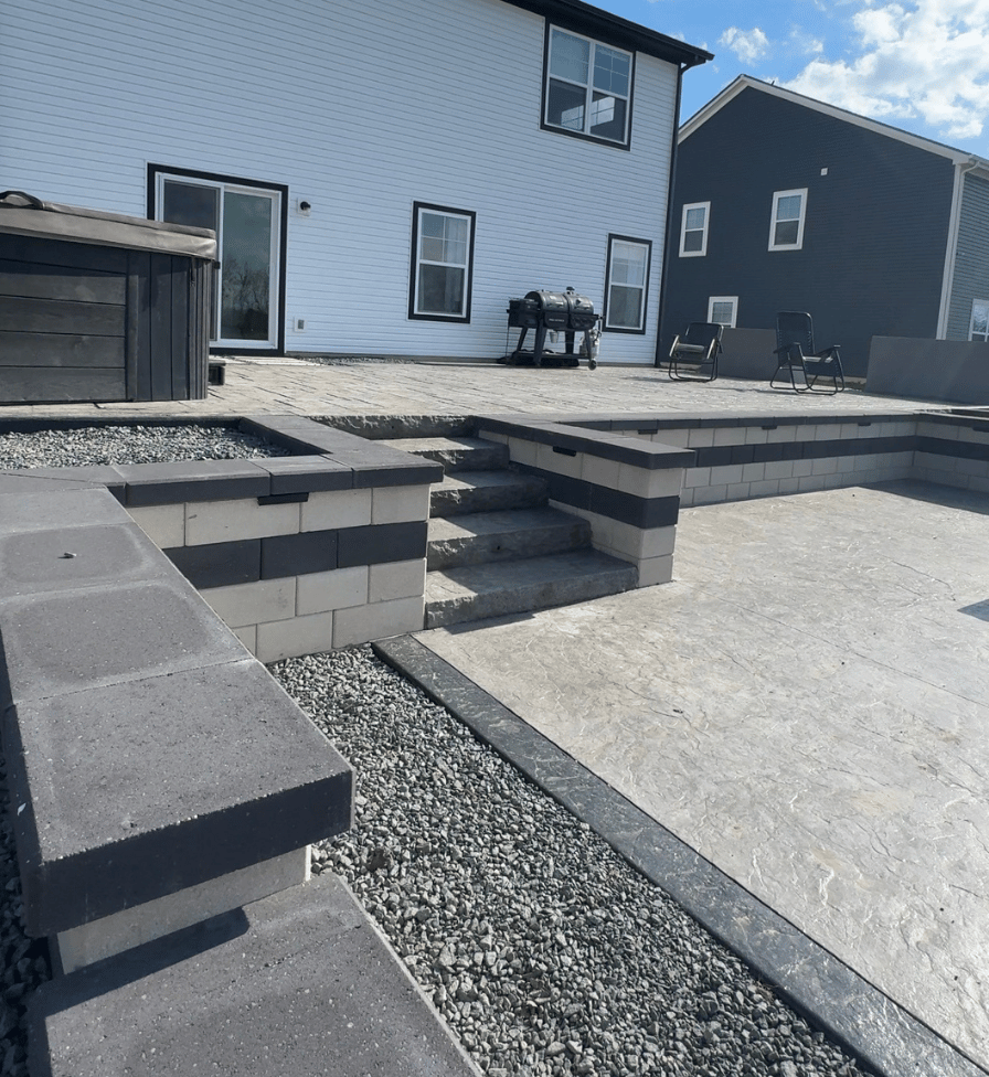 Modern backyard patio featuring stamped concrete flooring with textured pattern, tiered stone steps leading up to a raised deck area with two black chairs and a barbecue grill, bordered by walls with dark and light stone blocks, and gravel beds alongside the patio edges.