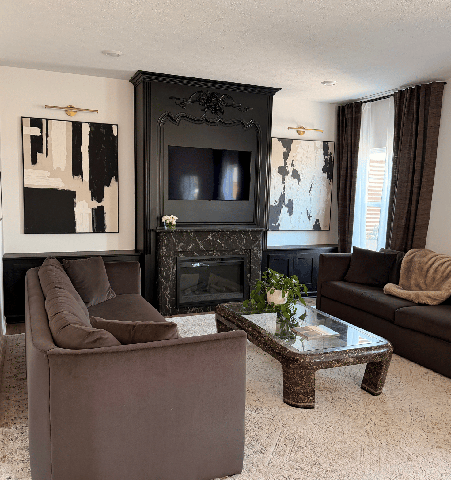Modern living room with two brown sofas facing each other across a rectangular marble and glass coffee table, a black ornate fireplace with a mounted TV above, abstract black and white paintings on either side of the fireplace, and large windows with brown curtains.