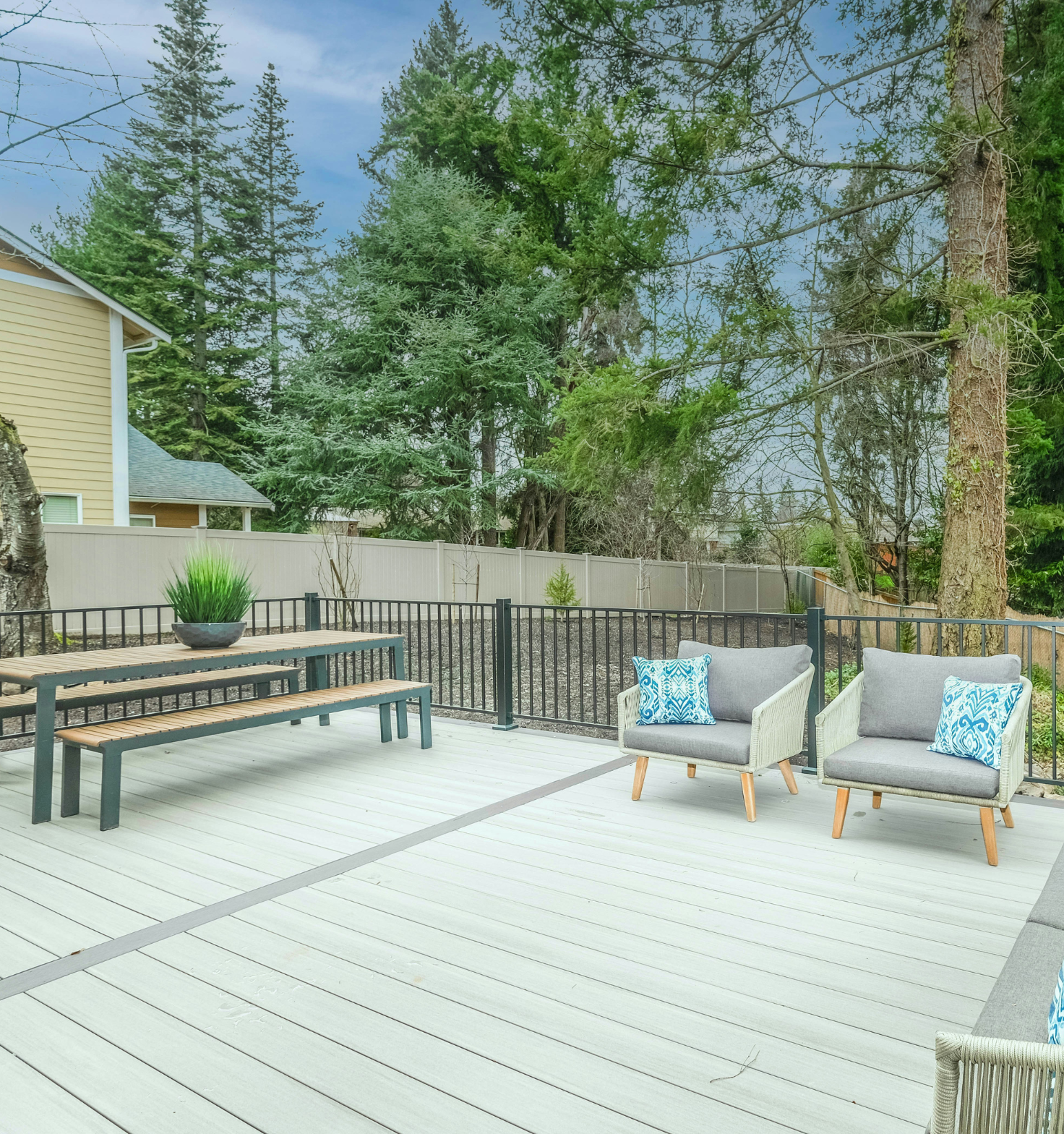 Outdoor deck area with light gray composite decking, a wooden picnic table with benches and a potted green plant on top, two cushioned armchairs with blue patterned throw pillows, black metal railing, and trees and a house in the background.