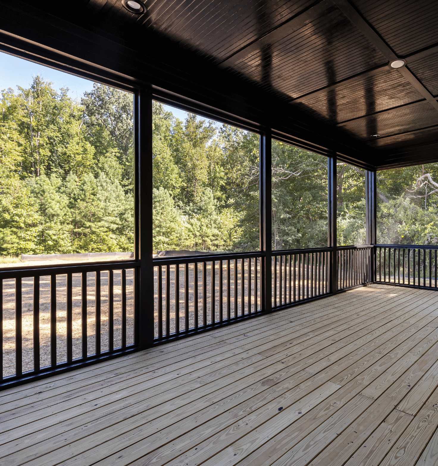 Spacious screened porch with light wooden floorboards, black railings, and a black paneled ceiling, overlooking a wooded area with green trees.