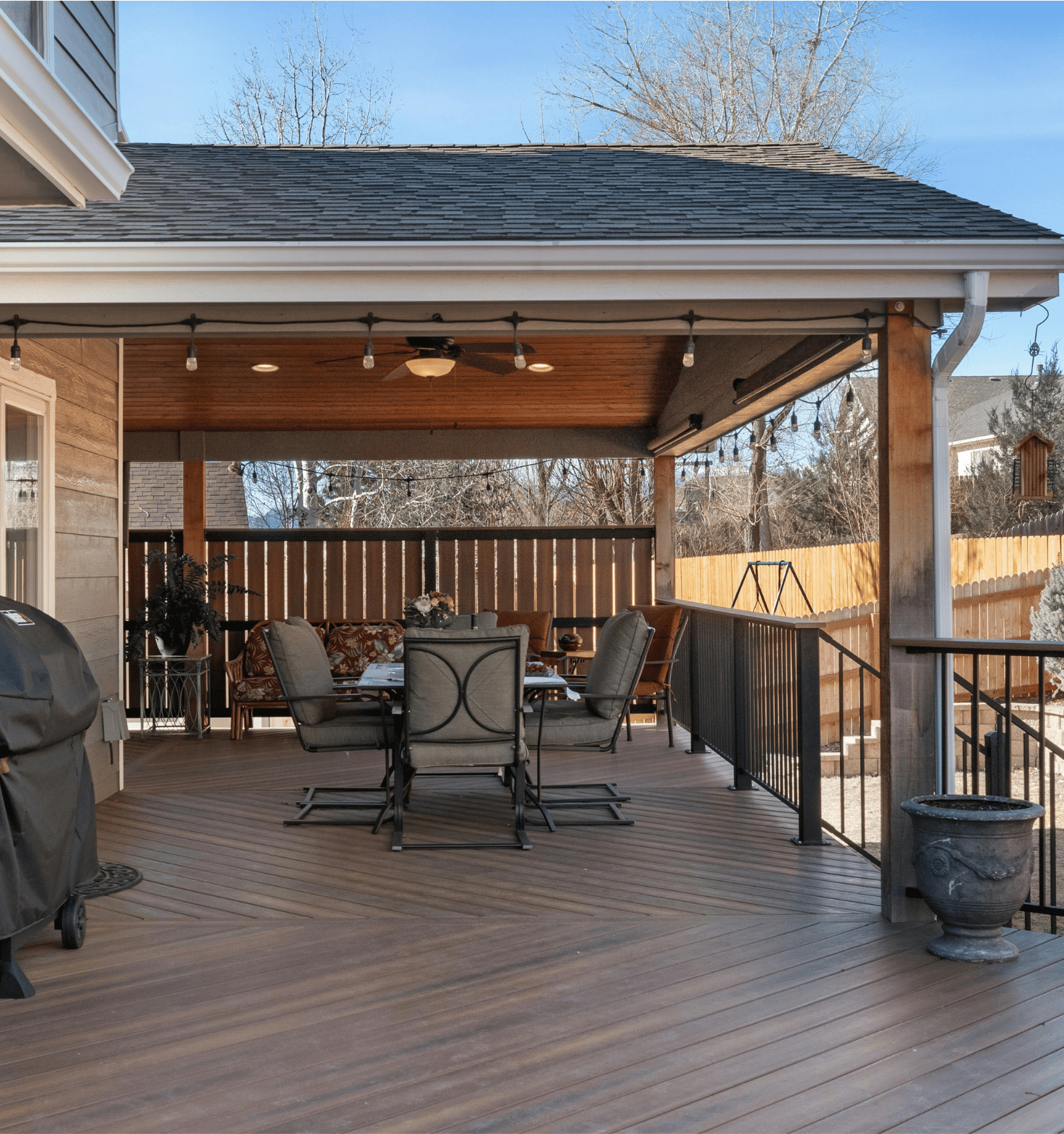 Covered outdoor patio with wood flooring and ceiling, featuring a dining table with cushions, a bench with floral cushions, string lights, ceiling fan, potted plants, and a grill on the left side.
