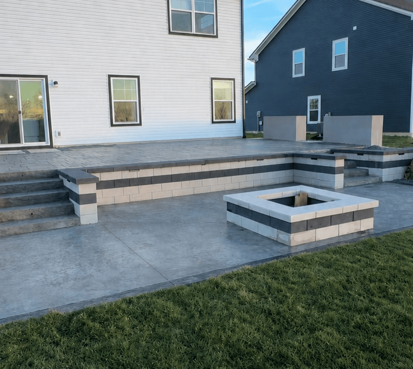Modern backyard patio with stamped concrete flooring, featuring a raised seating area with two steps on either side, and a square fire pit bordered by light and dark gray concrete blocks, adjacent to a grassy lawn and two houses in the background.