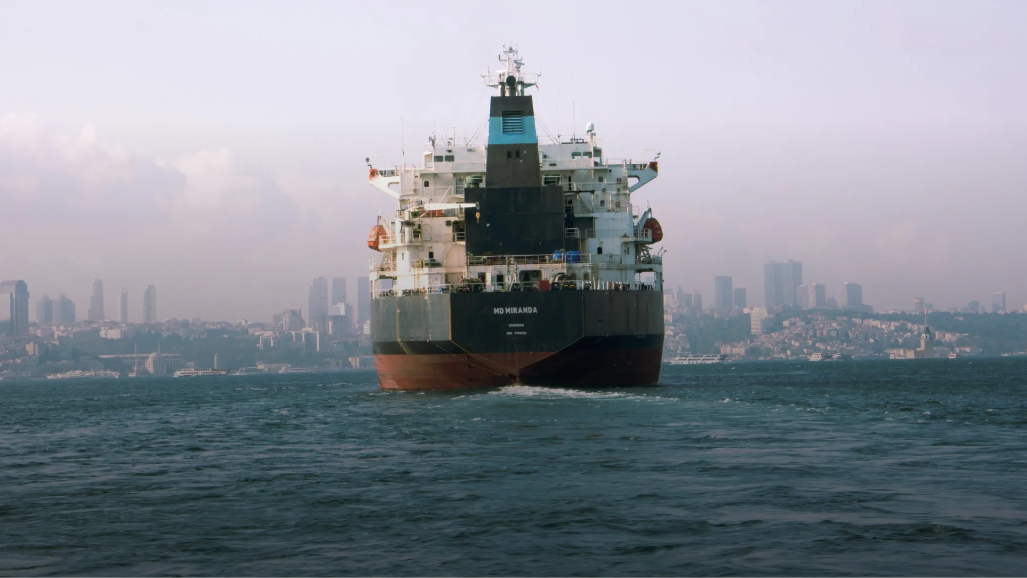 Large cargo ship named MD Miranda on water with city skyline and buildings in the background.