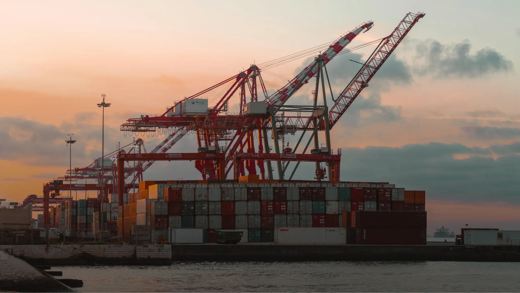 Shipping containers stacked at a port with large cranes against a sunset sky.