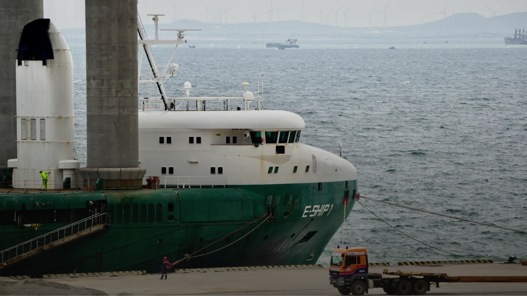 Large green and white ship docked with two workers and a flatbed truck nearby at the harbor.