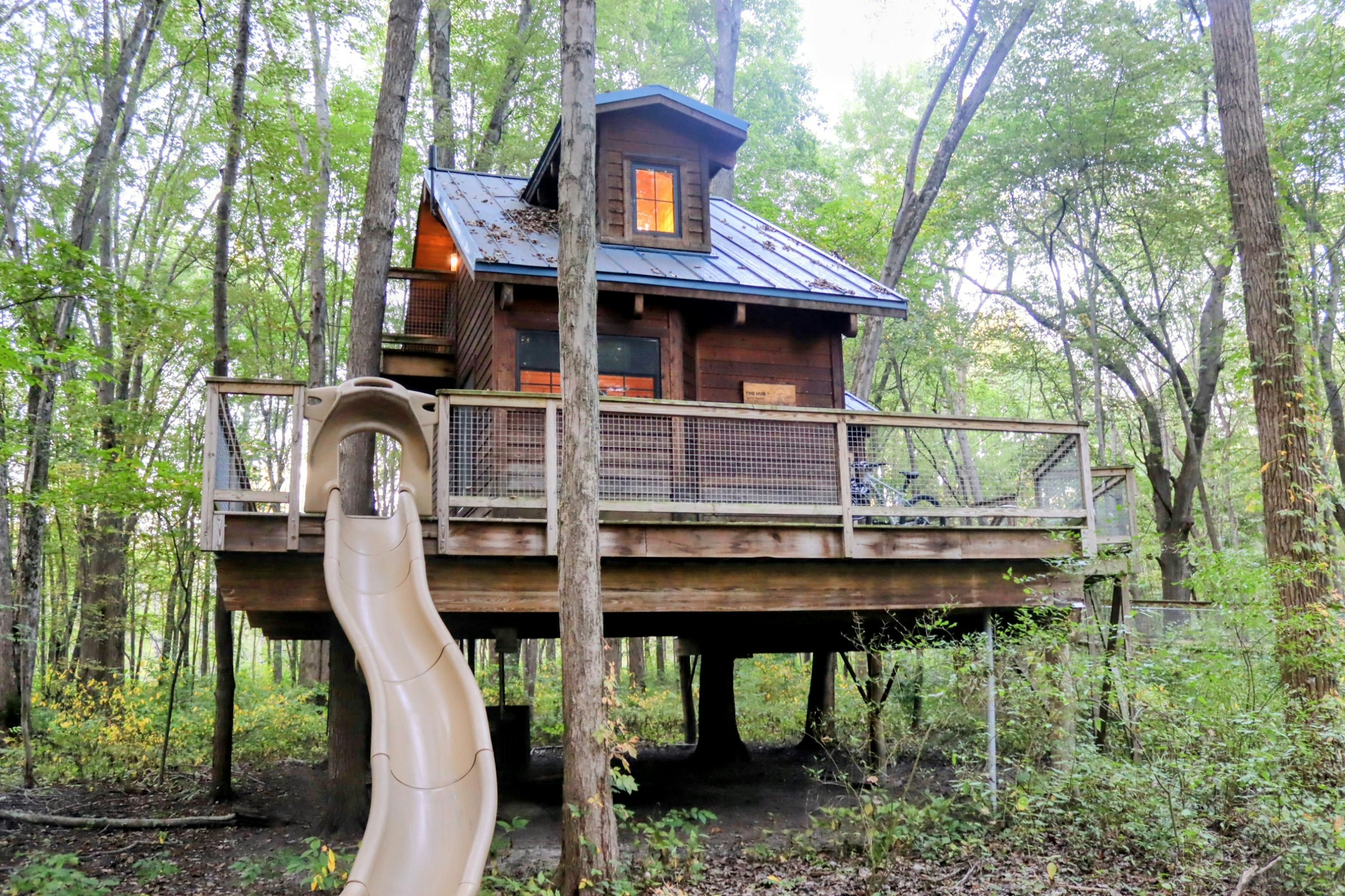 Cabane perchée en bois sur pilotis au milieu des arbres, avec terrasse, garde-corps grillagé, toiture métallique et toboggan